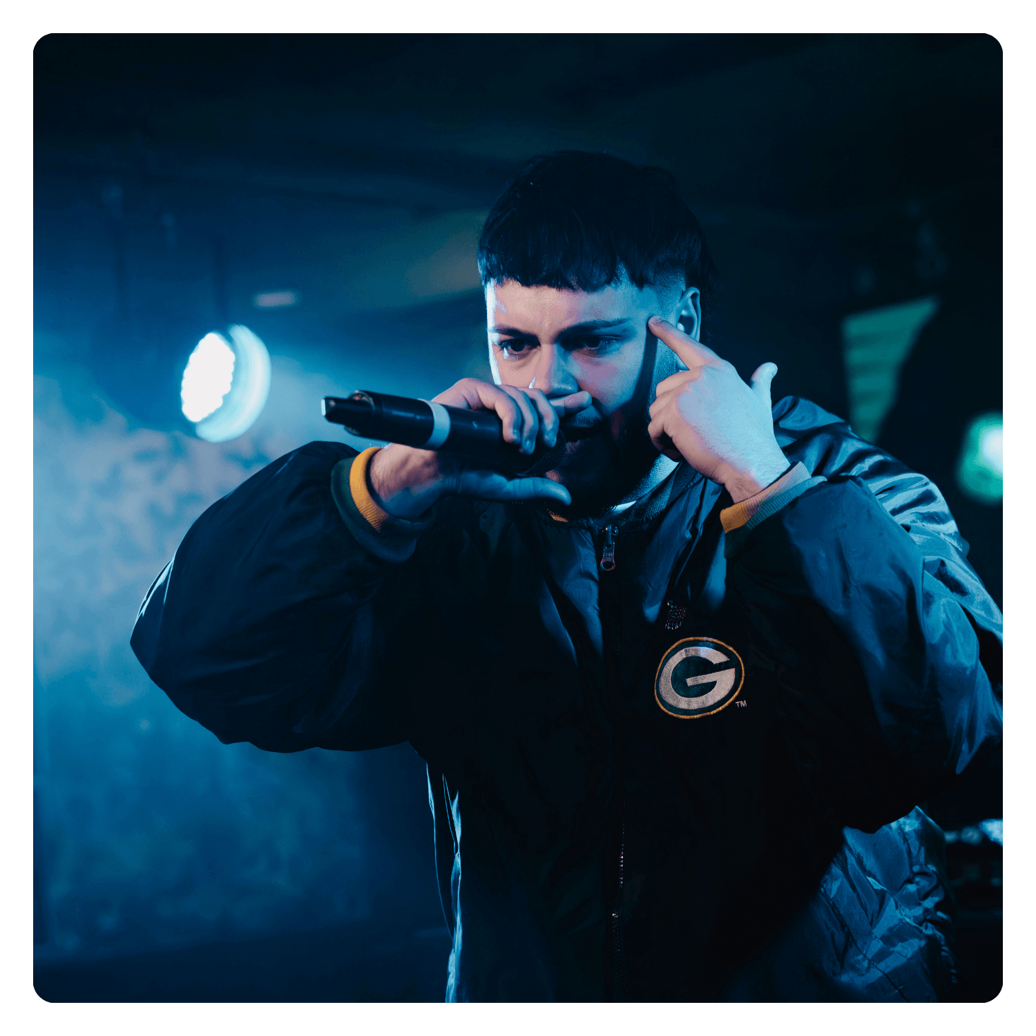 A man performing on stage with a microphone, wearing a black jacket with a sports team logo, and pointing to his head with his finger, under blue stage lighting.