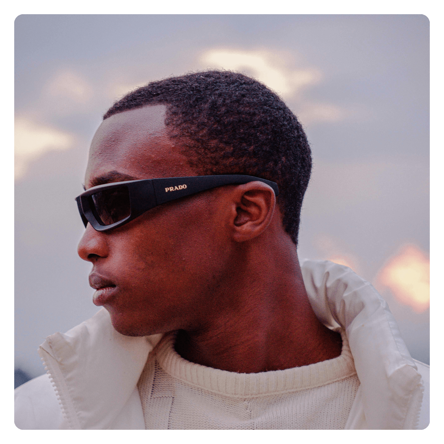 A young man with dark skin wearing black Prada sunglasses and a white puffer jacket, looking to the left against a cloudy sky background.