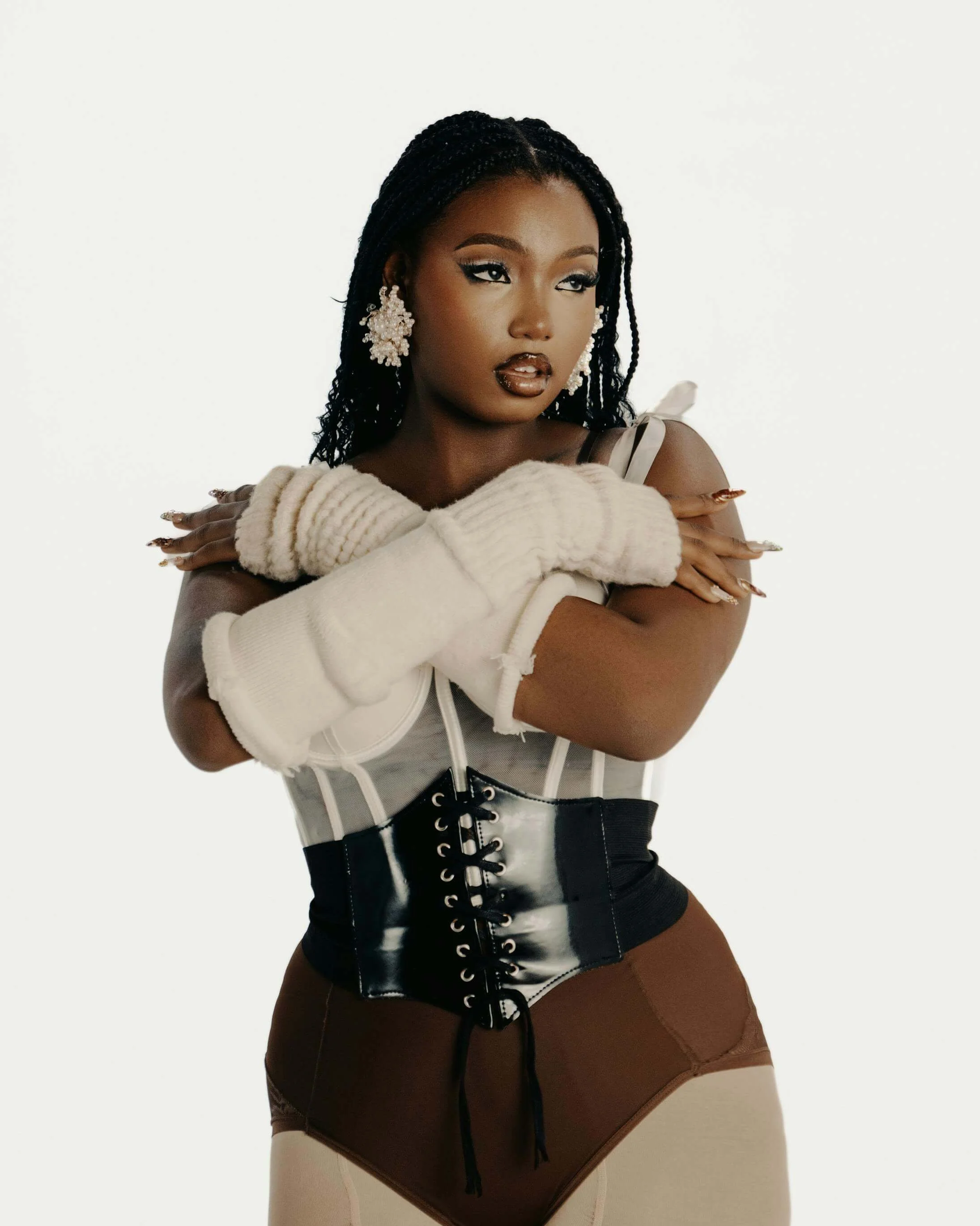 A woman with dark skin and braided hair, wearing large floral earrings and makeup, crosses her arms over her chest and gazes to the side. She has on cream-colored gloves, a white corset-style top, a black shiny waist cincher, and brown high-waisted bottoms.