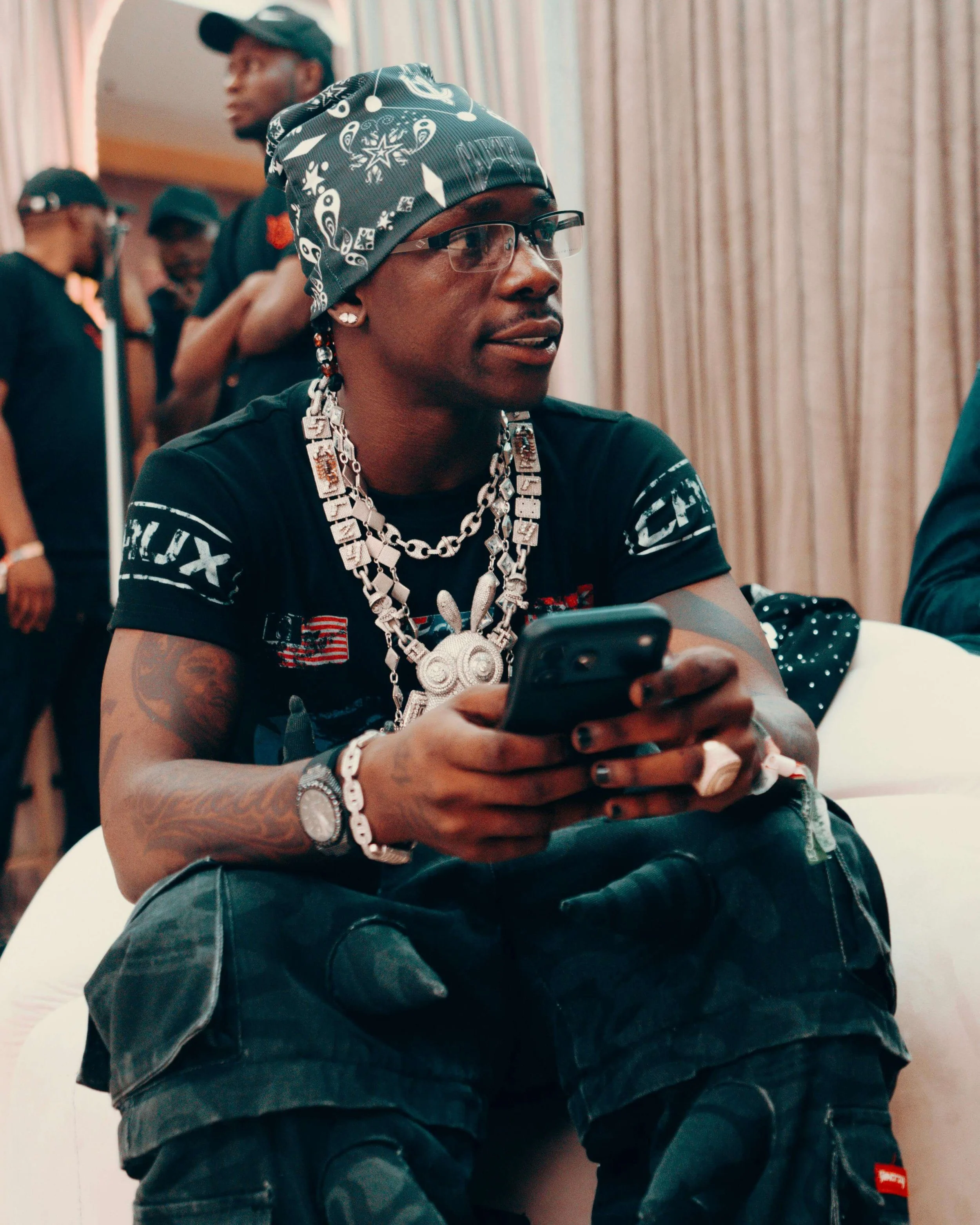 A man wearing a bandana, glasses, and multiple silver necklaces is sitting while holding a phone. There are other people in the background at an indoor event.