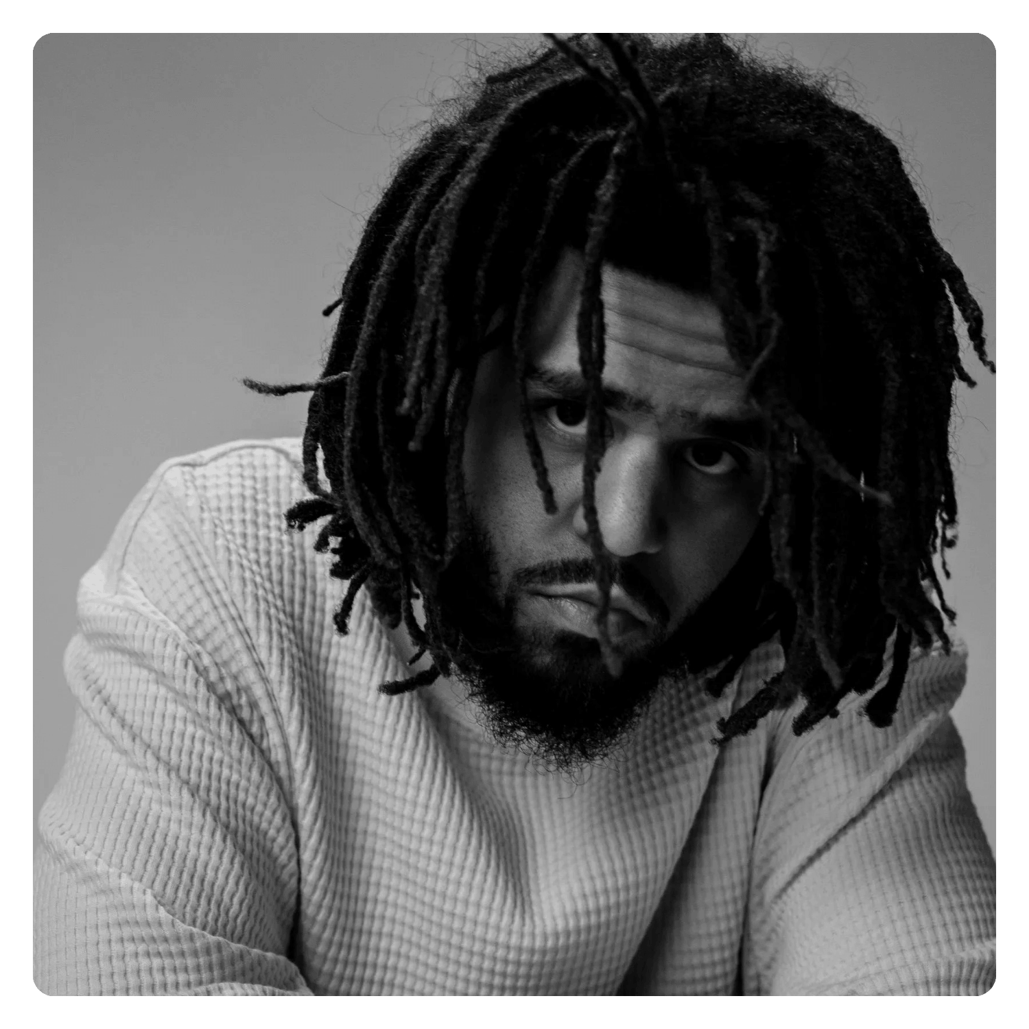 Black and white portrait of a man with dreadlocks and a beard, wearing a textured sweater, looking directly at the camera with an intense expression.