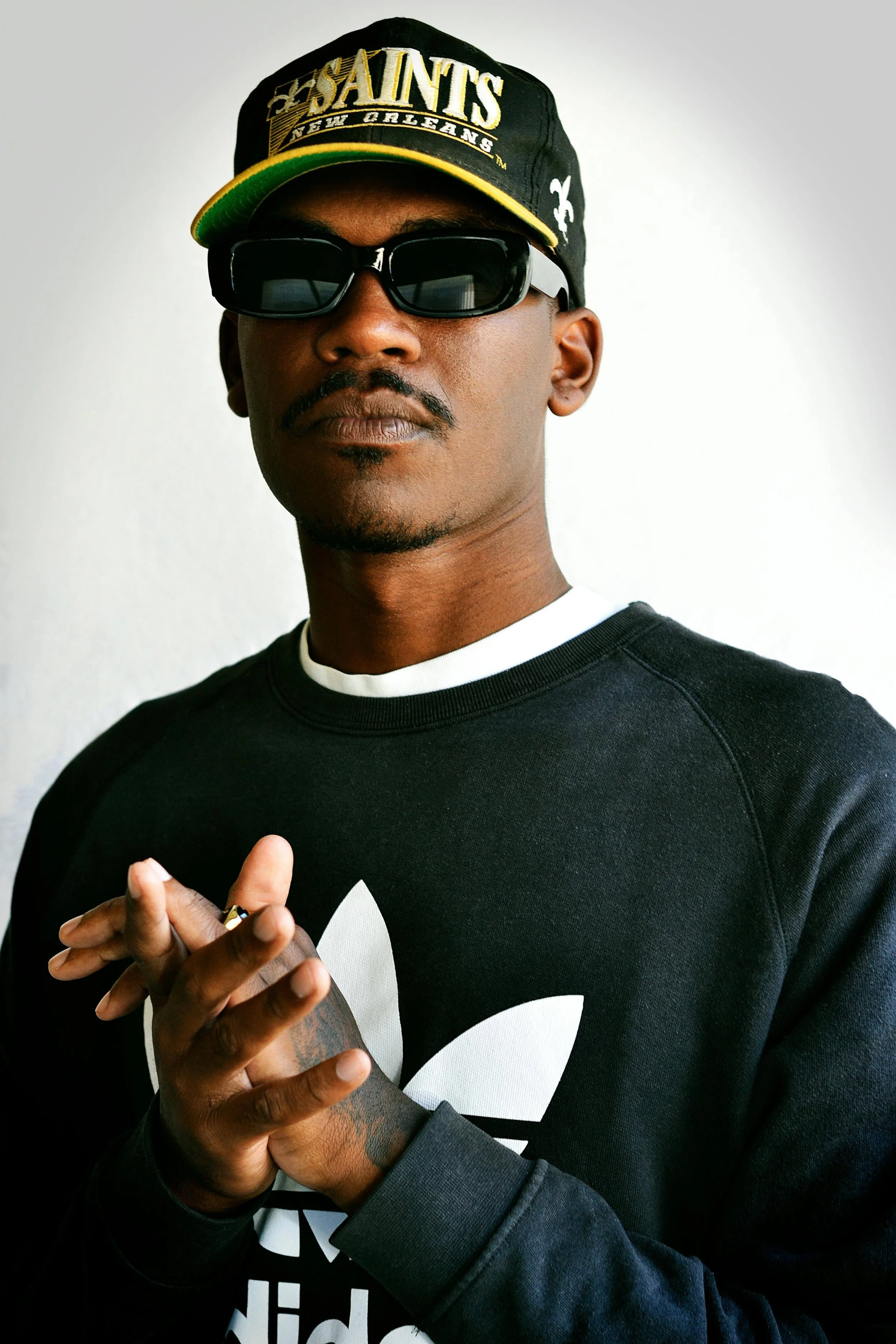 A man wearing a black New Orleans Saints cap, black sunglasses, and a black Adidas sweatshirt with the white trefoil logo. He has a mustache and goatee, and is holding his hands together in front of him.