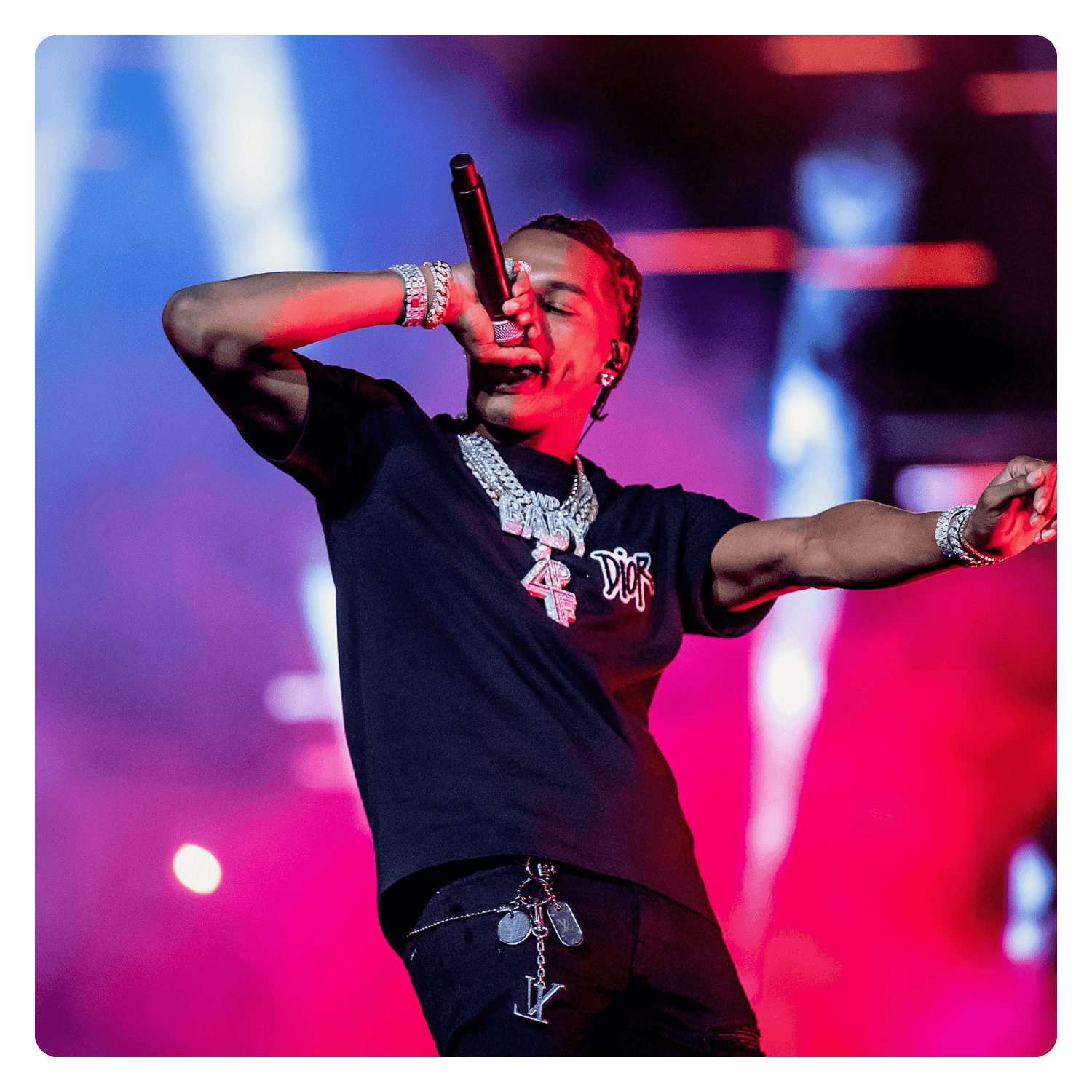 Rapper performing on stage wearing jewelry and black clothing, holding microphone, with colorful lights in the background.