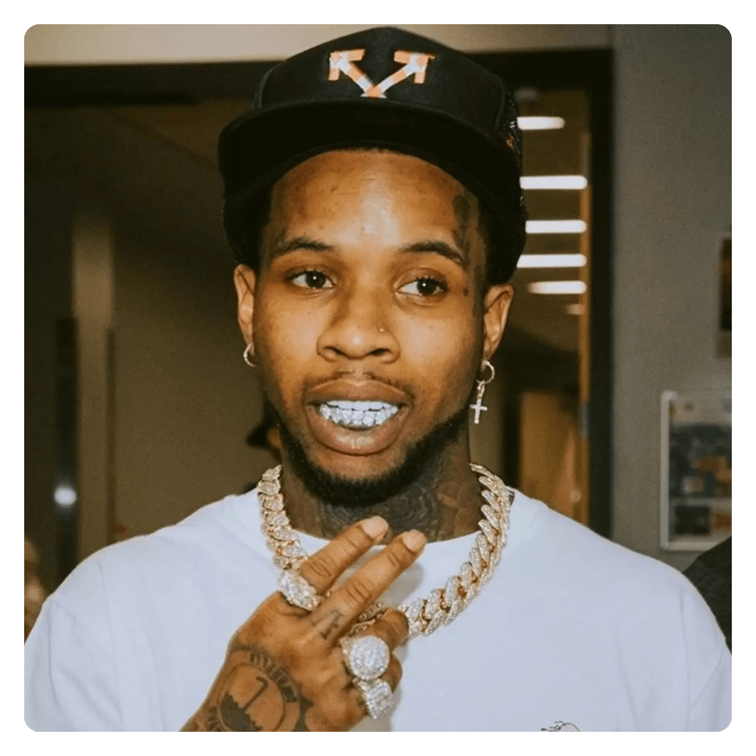 Young Black man with tattoos, wearing a black cap, diamond jewelry, and a white shirt, making a gesture with his hand near his face in an indoor setting.
