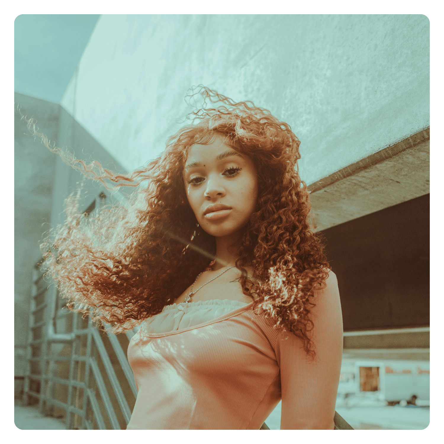Young woman with long, curly hair standing outdoors under a concrete structure with buildings in the background.