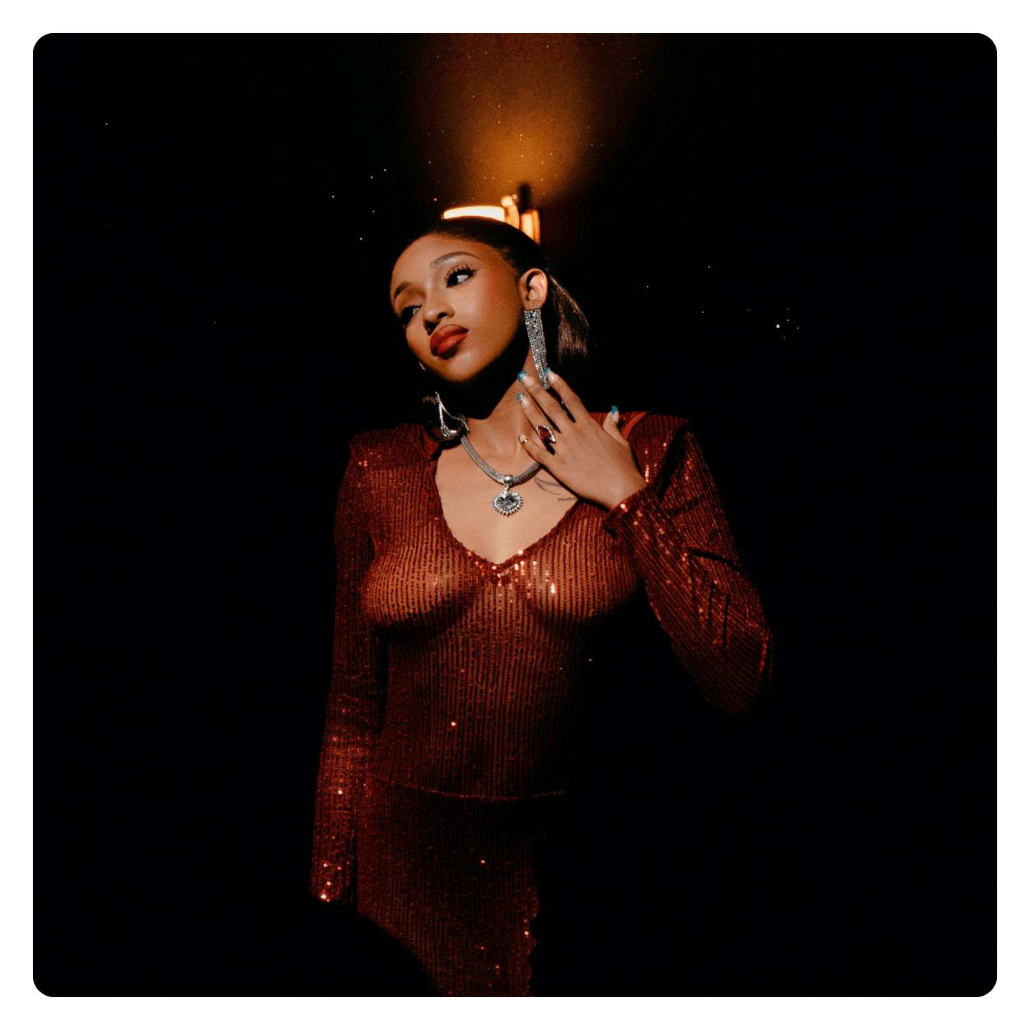 A woman dressed in a shimmering red dress with jewelry, posing against a dark background with a light above her head.