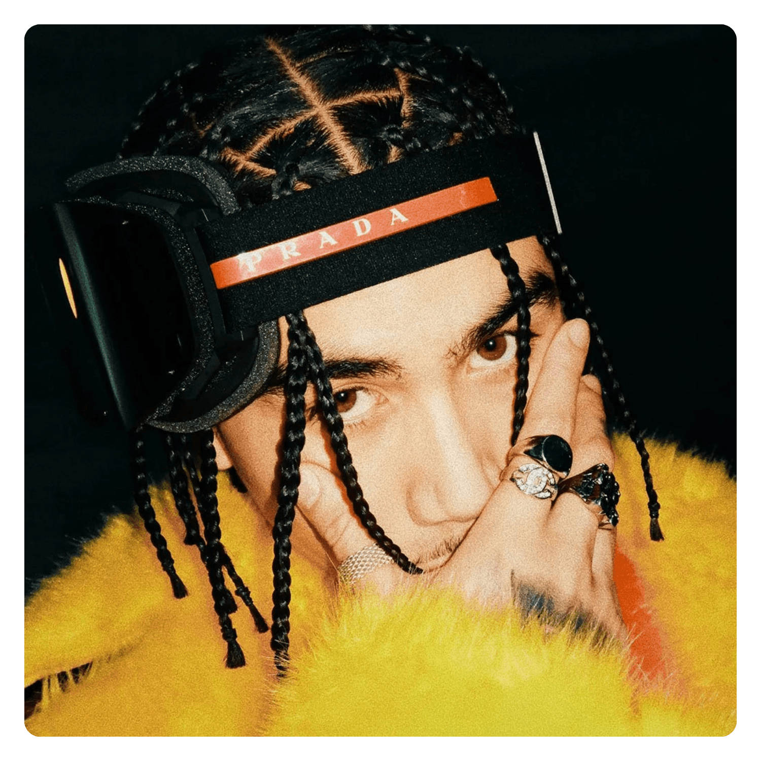 A person with braided hair, wearing a yellow fuzzy coat, jewelry including rings and a bracelet, and a black Prada headband, poses with their fingers near their face against a black background.