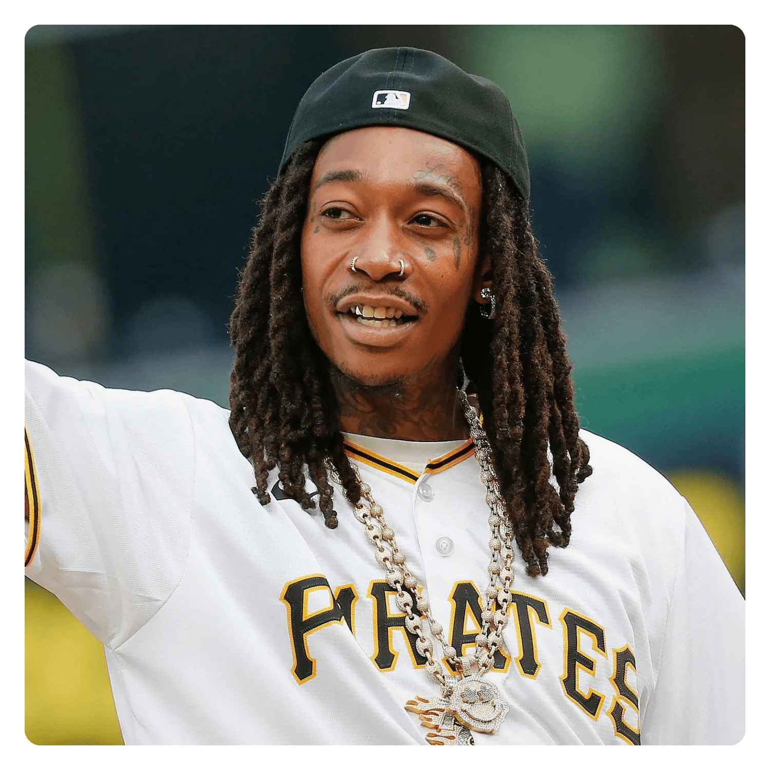 A man with dreadlocks wearing a black baseball cap, a white Pittsburgh Pirates jersey, and jewelry, smiling and raising his arm, outdoors with blurred background.
