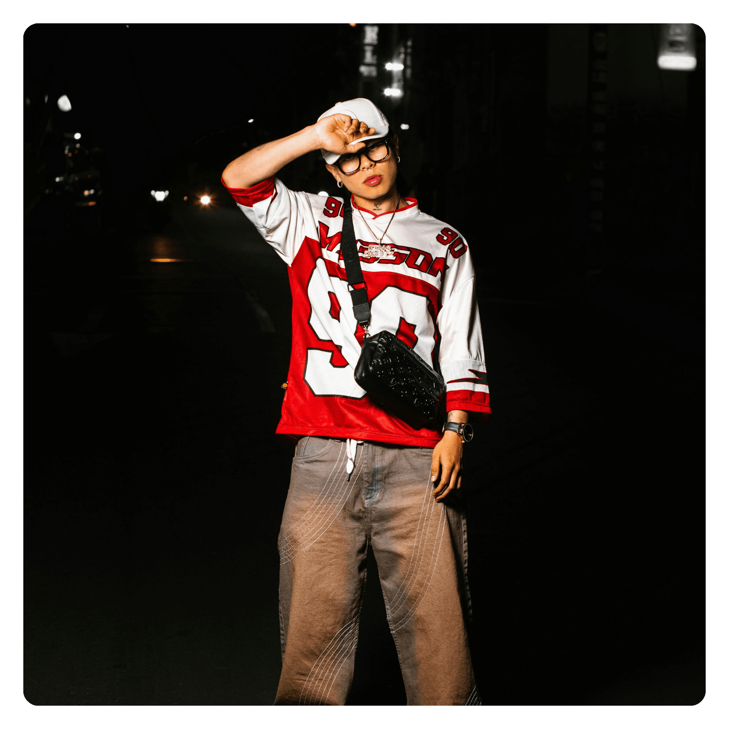 Person standing on street at night, wearing a red and white sports jersey, beige pants, black glasses, white cap, carrying a small black shoulder bag, with streetlights in the background.