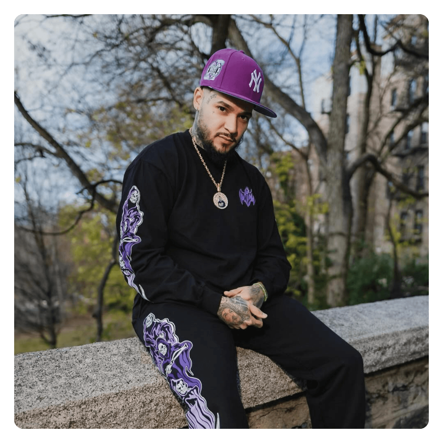 A man with tattoos on his hands and neck, wearing a black long sleeve shirt with purple designs, black pants with purple designs, a purple cap, and a gold chain with a medallion, sitting on a stone ledge outdoors with trees and buildings in the backg