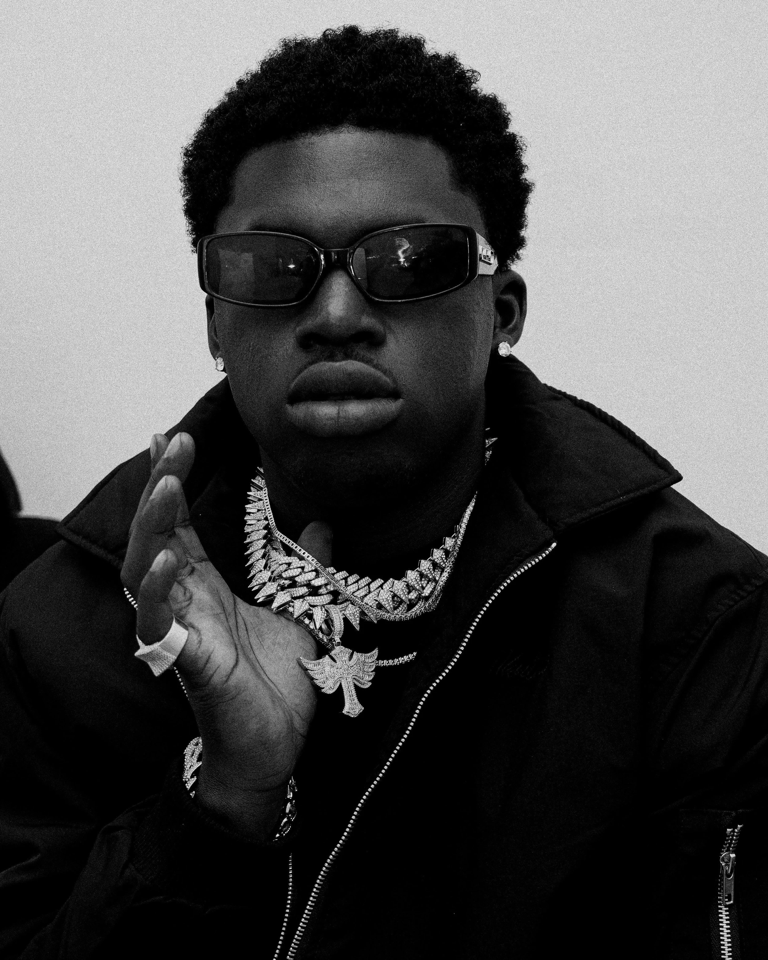 A young man wearing dark sunglasses, jewelry including a chain necklace, a cross pendant, and rings, dressed in a dark jacket, in black and white photo.
