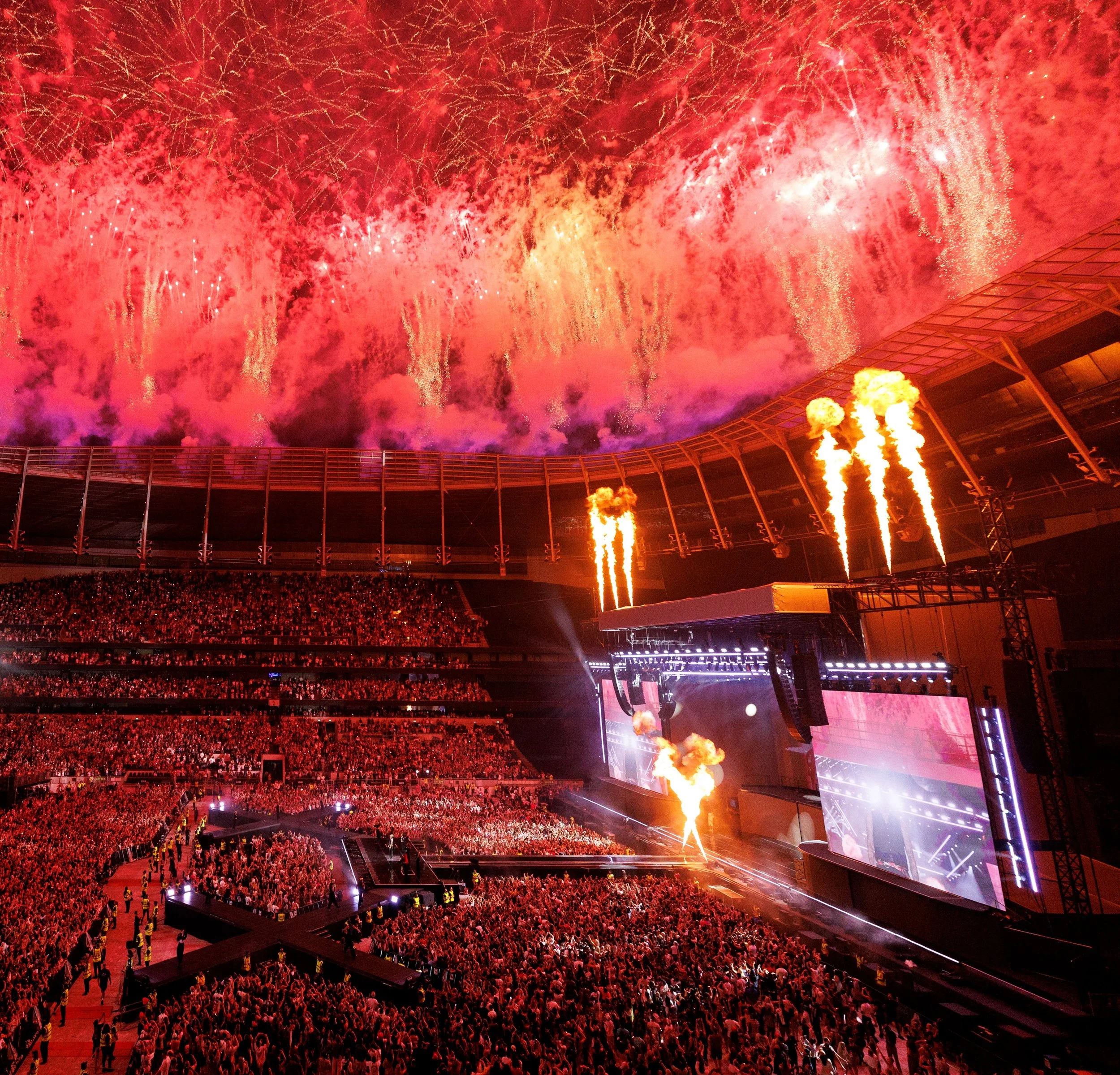 Fireworks display over a packed stadium during a concert or event, with bright explosions in the sky, stage lights, and pyrotechnics.