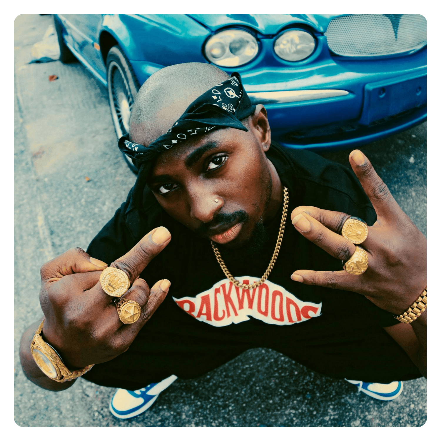 A man with a shaved head and dark skin is squatting on the ground in front of a blue car. He is wearing a black T-shirt with red and white lettering, multiple gold rings, a gold watch, a gold chain, and has a black bandana around his head. He is maki