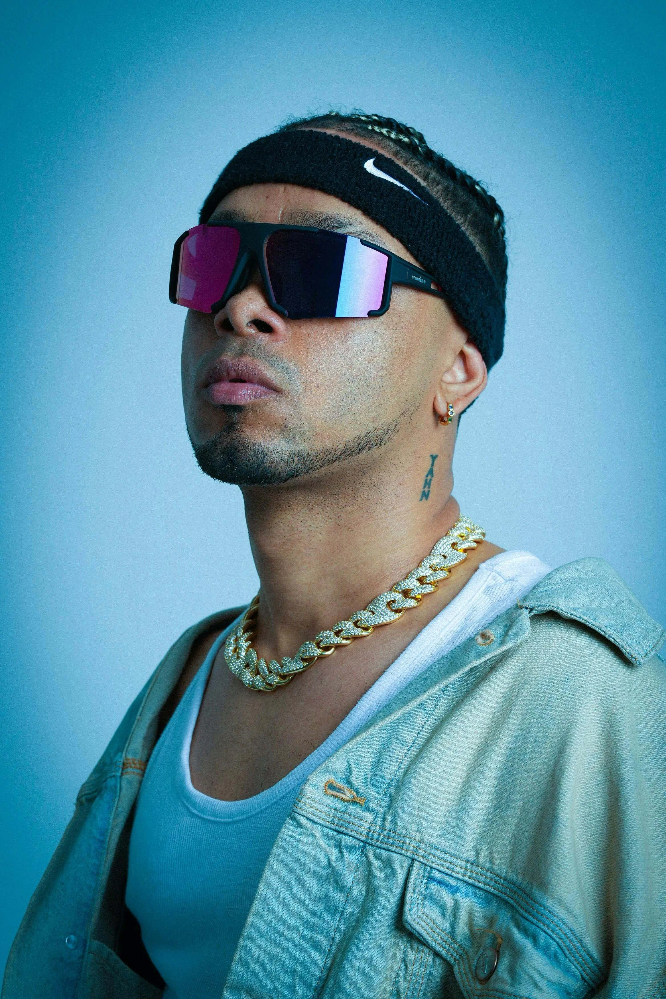 A man wearing reflective sunglasses, a black Nike headband, a gold chain necklace, and a white shirt under a light green jacket, with a tattoo on his neck and a small earring.