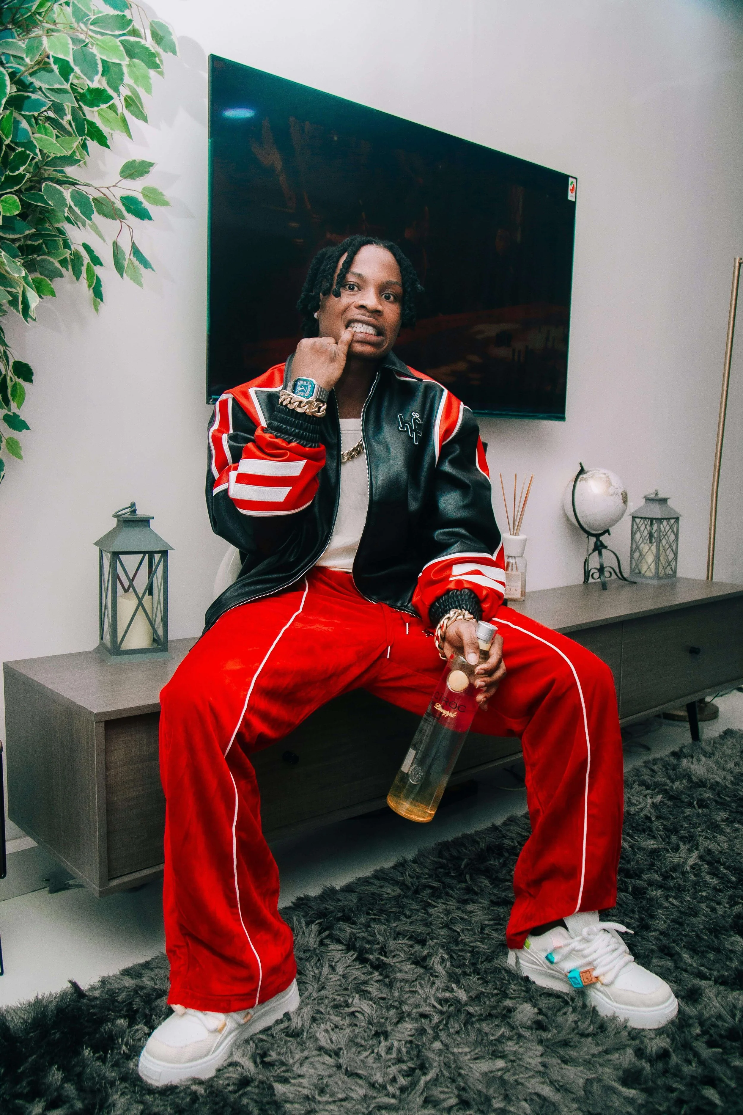 A man sitting on a wooden cabinet, holding a bottle of what appears to be alcohol, in a modern living room with a large TV, decorative lanterns, a small globe, a plant, and a black fluffy rug.