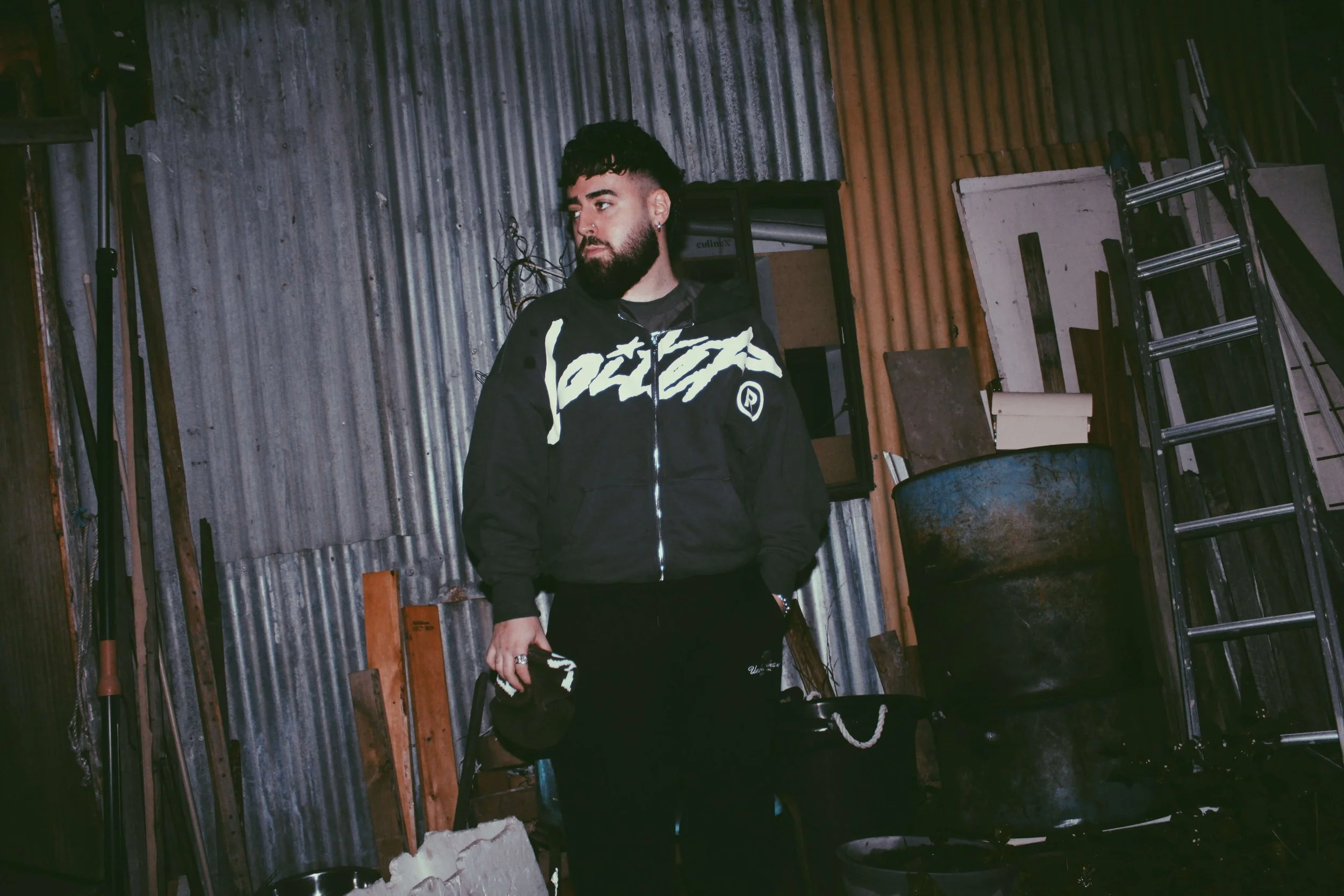 A young man with dark hair, beard, and earrings standing in a rustic, industrial space with corrugated metal walls, holding a black cap in his left hand, wearing a black jacket with white graphics, and black pants.