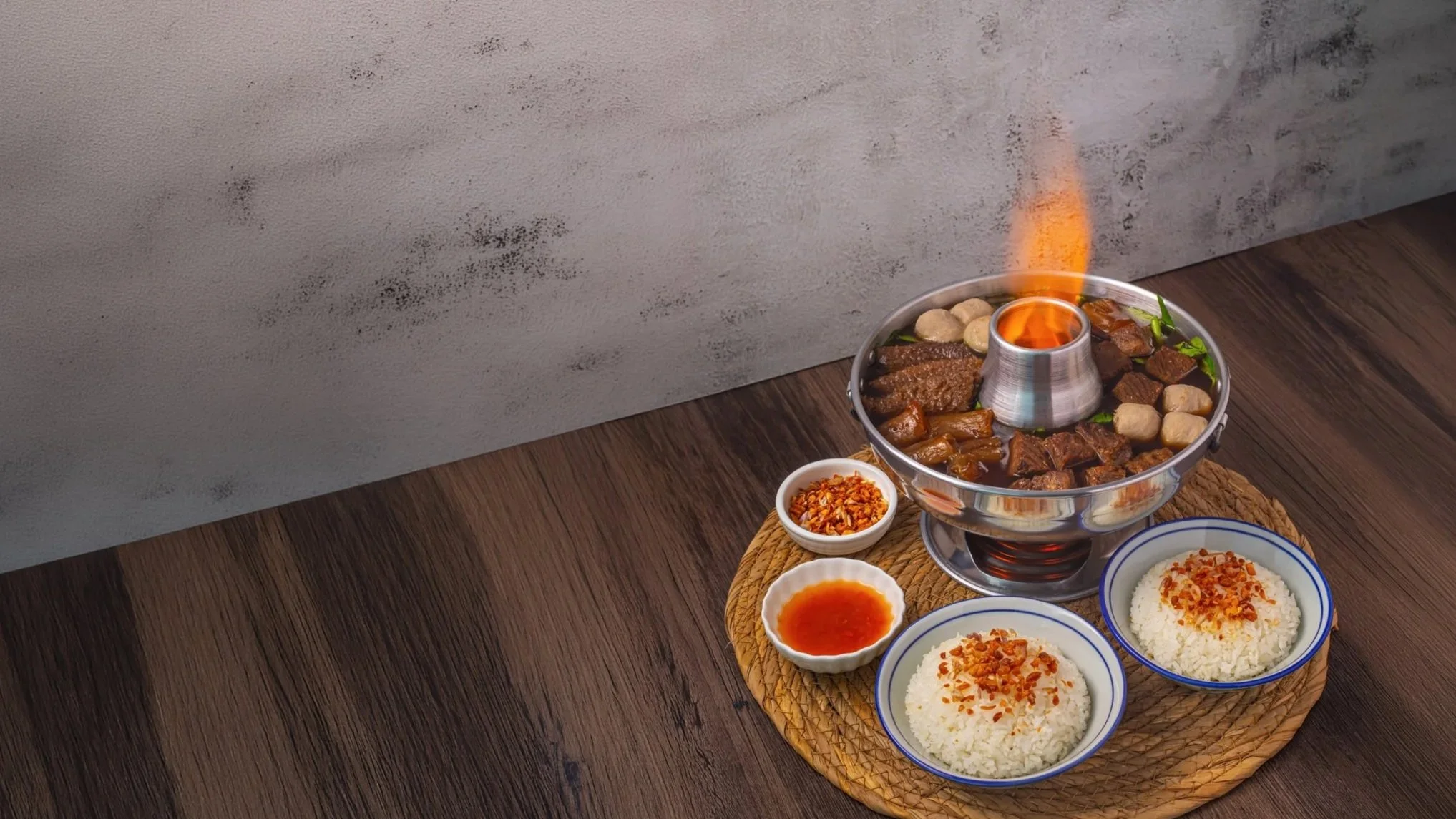 A thai firepot with spicy chilli sauce, and bowls of rice garnished with fried garlic, arranged on a woven placemat on a wooden table.