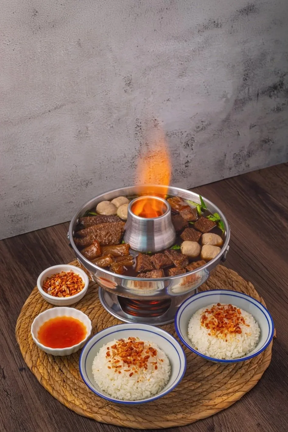 A hot pot with various meats and vegetables, surrounded by bowls of rice, sauces, and condiments. The hot pot has a central chimney with a small flame, and the food inside includes beef, meatballs, and greens.