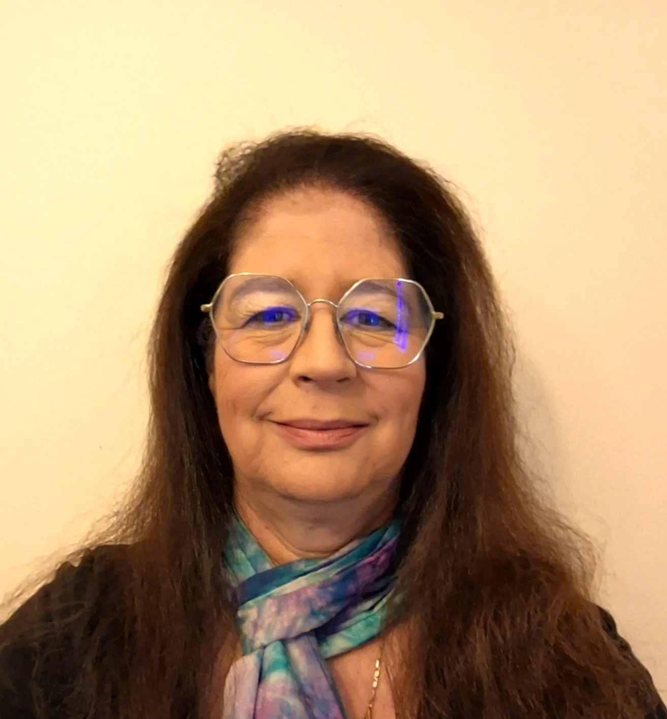 A woman with long, wavy brown hair wearing large, round glasses with a slight tint, smiling in front of a plain light-colored wall. She has on a multicolored scarf and a dark top.