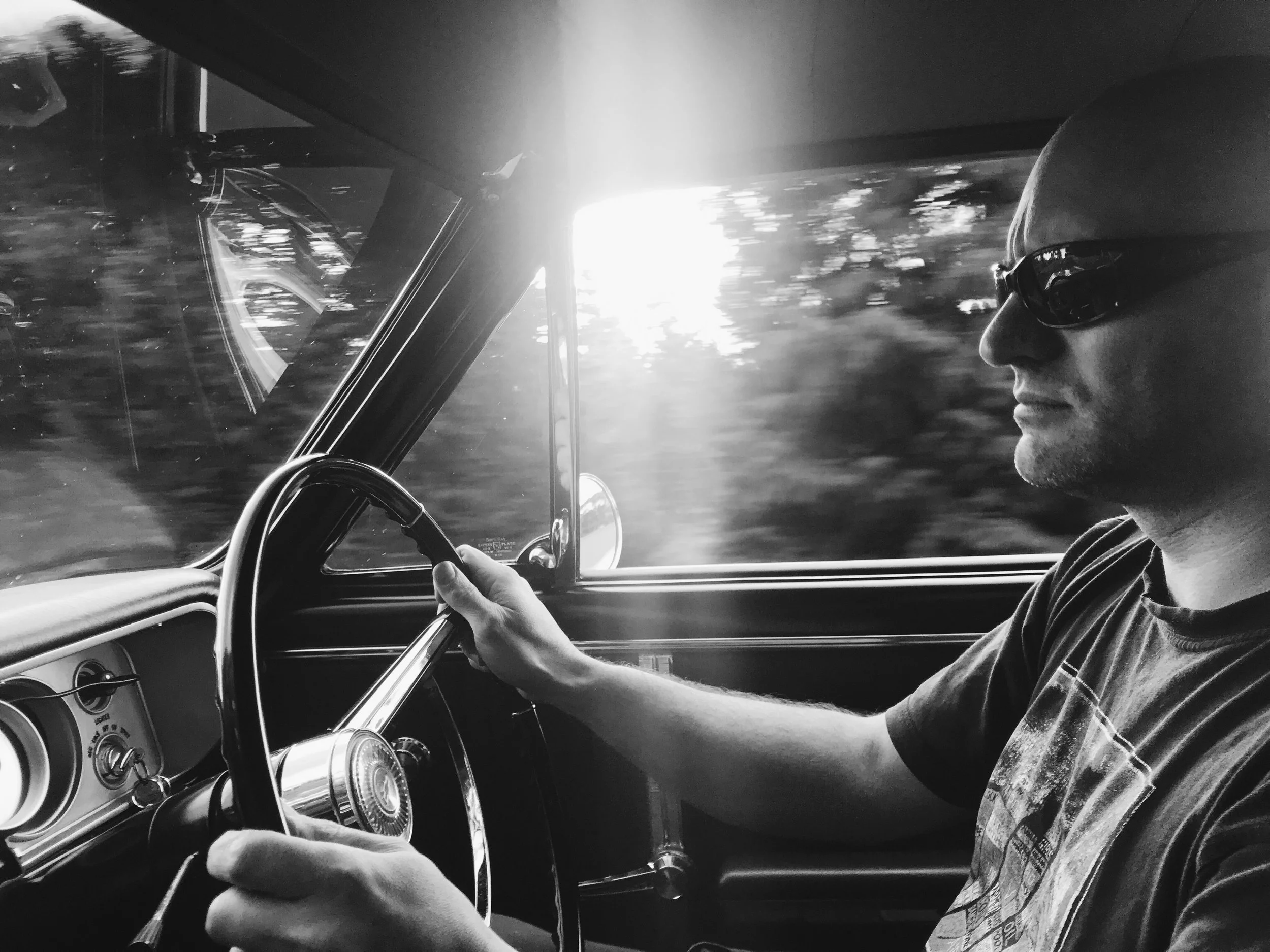 A man wearing sunglasses and a plaid shirt driving a vintage car on a sunny day.