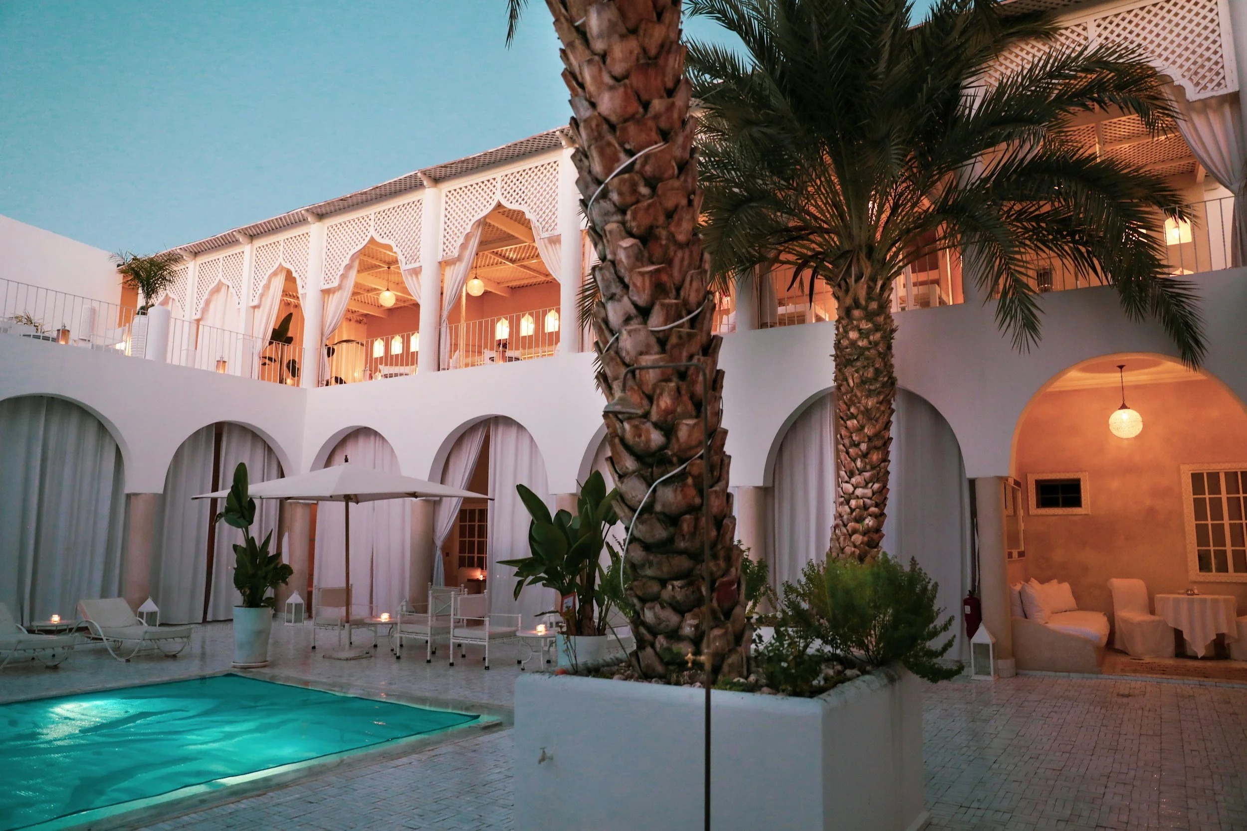 Cour intérieure d'hôtel avec piscine, palmiers, zones de détente et bâtiments blancs lumineux sous un ciel bleu soir.