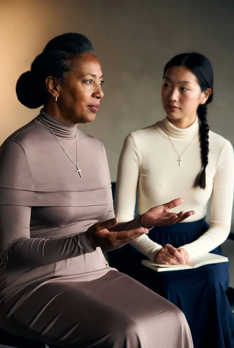 An older woman and a young woman sitting and having a conversation, both wearing cross necklaces, with the older woman gesturing with her hands.