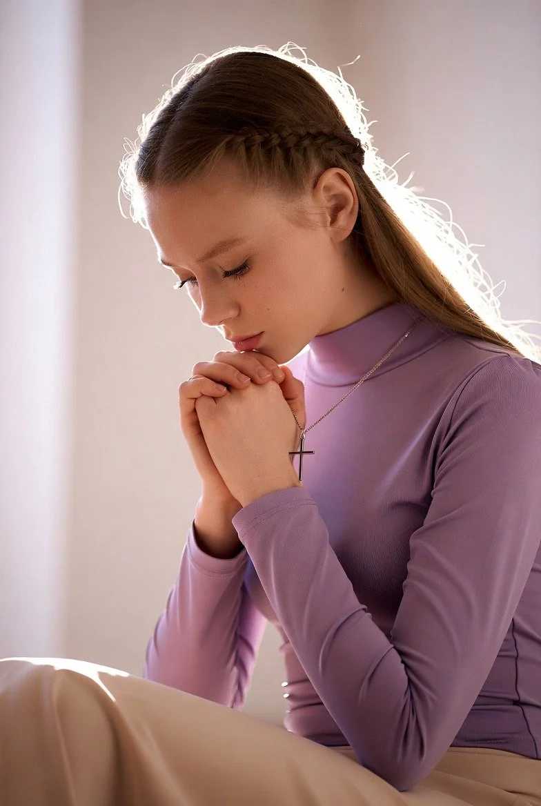 A young woman praying with her hands clasped and eyes closed, wearing a lavender long-sleeve top and a cross necklace, with sunlight backlighting her hair.