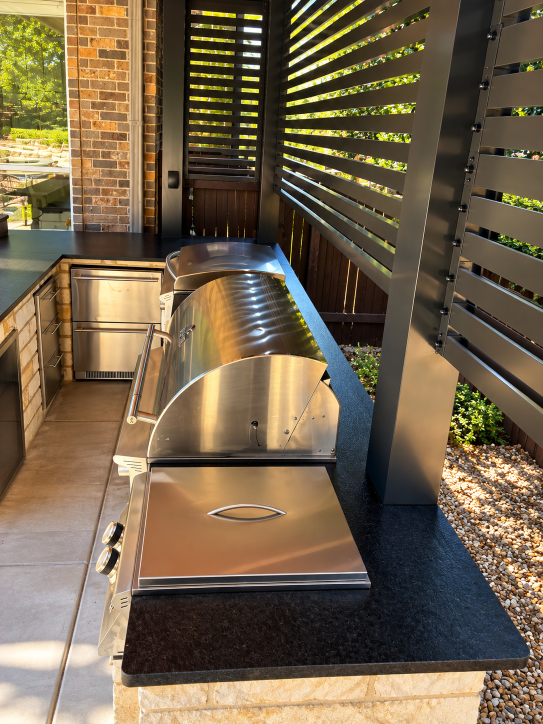 Outdoor kitchen with stainless steel grill, black countertop, brick wall, wooden privacy fence, and horizontal metal slats.