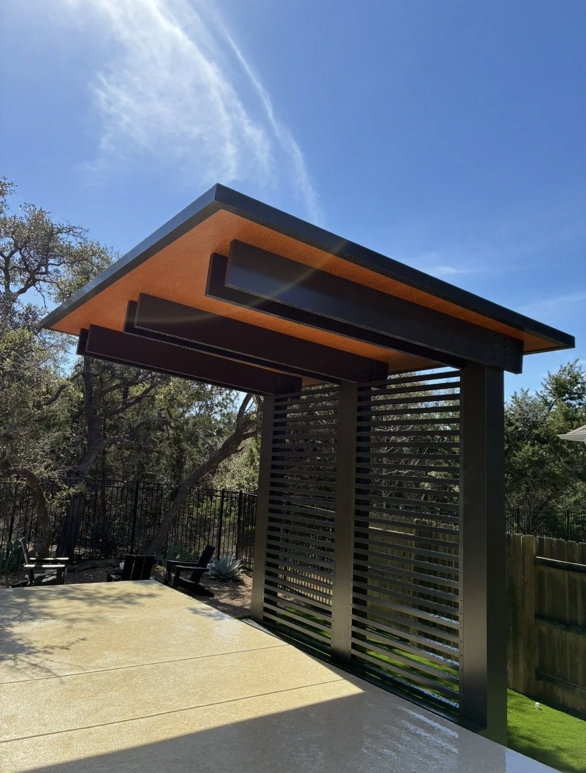 Modern outdoor structure with orange roof, black beams, and slatted metal wall, set in a landscaped backyard with trees and a concrete patio.
