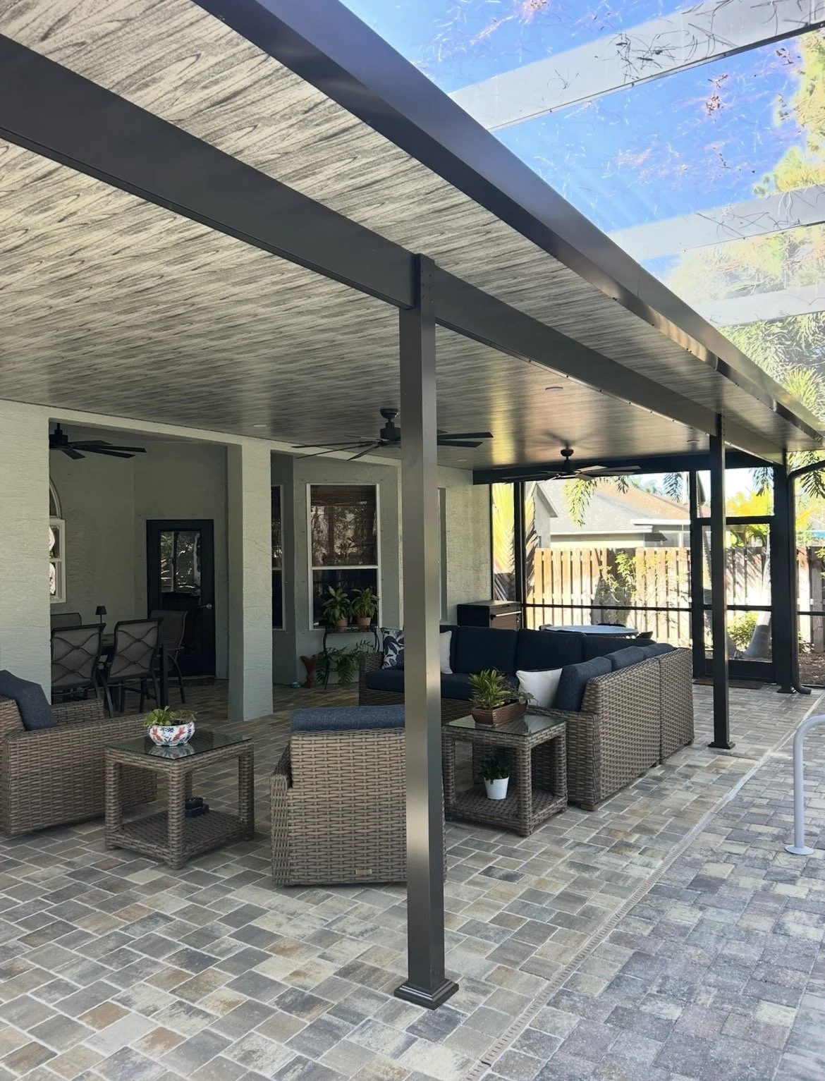 Covered outdoor patio with wicker and cushioned seating, potted plants, and a tiled floor, adjacent to a house with screened windows and a patio door.