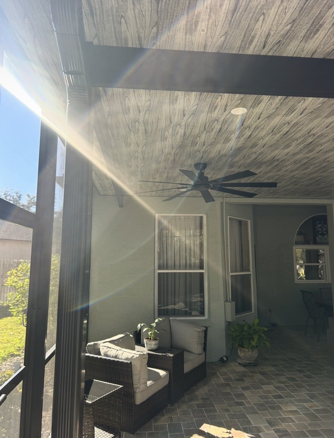 Sunlit outdoor patio with wicker sofa, potted plants, ceiling fan, and large windows.