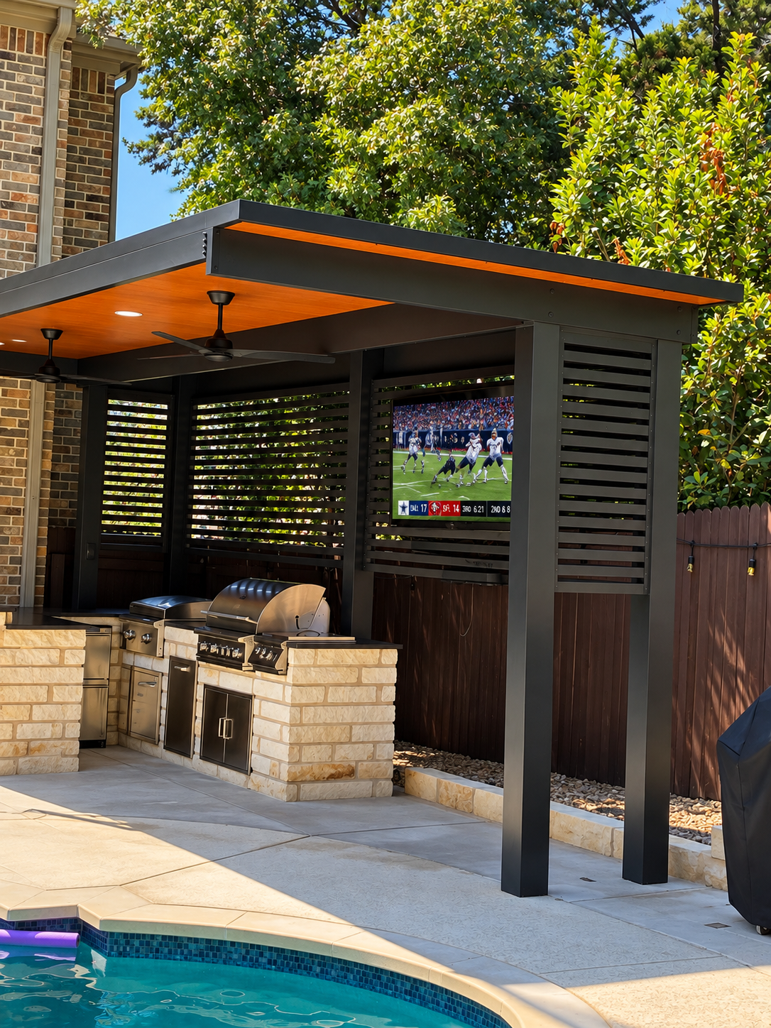 Outdoor patio area with a built-in grill station, countertop, and a mounted flat-screen TV showing a football game. There is a swimming pool in the foreground with a pool noodle floating on the water. The space is shaded by a modern pergola with ceil