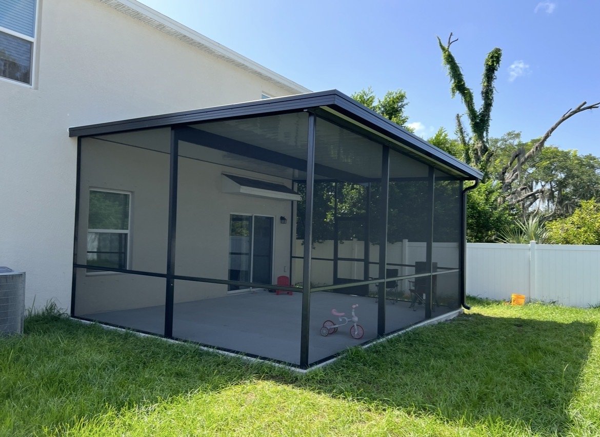 Backyard screened porch with a black frame, attached to a white house, with a green lawn and trees in the background.