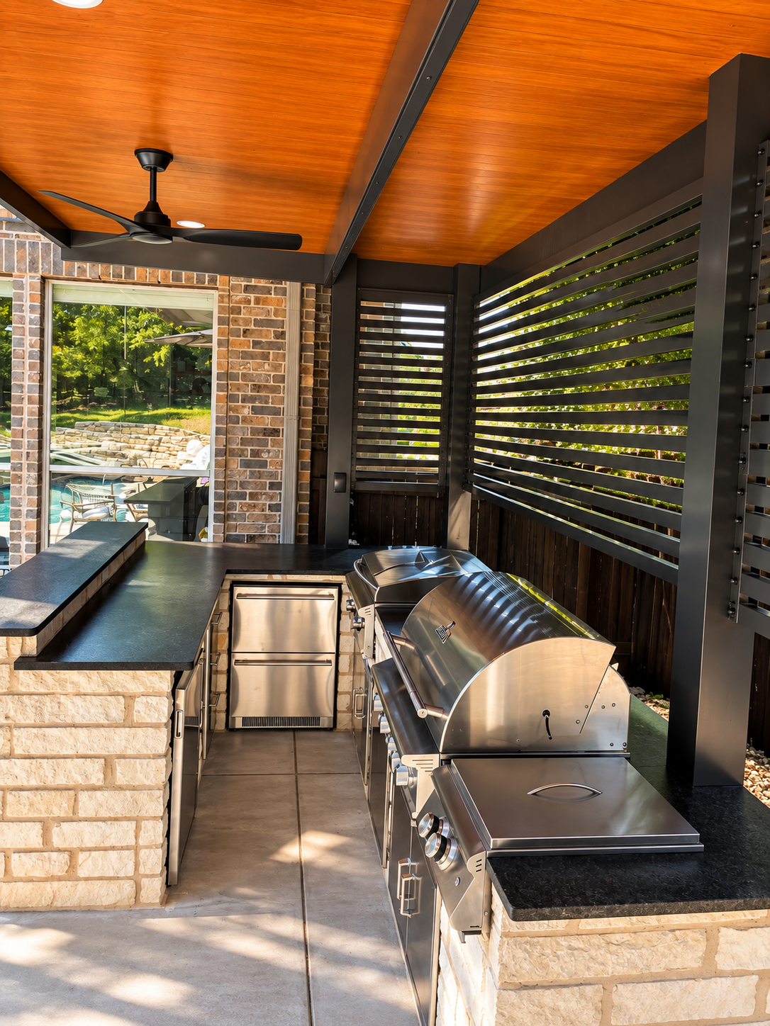 Outdoor kitchen with a stainless steel barbecue grill, black countertop, brick and black wood fencing, and a ceiling fan with an orange wood ceiling.