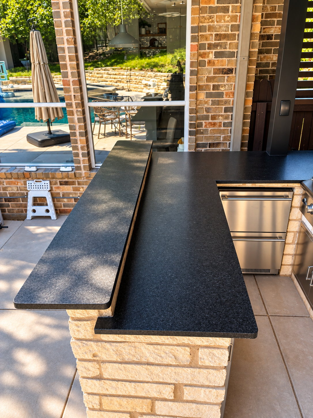Outdoor kitchen with black granite countertops, brick base, and stainless steel appliances, overlooking a backyard with a pool and patio furniture.