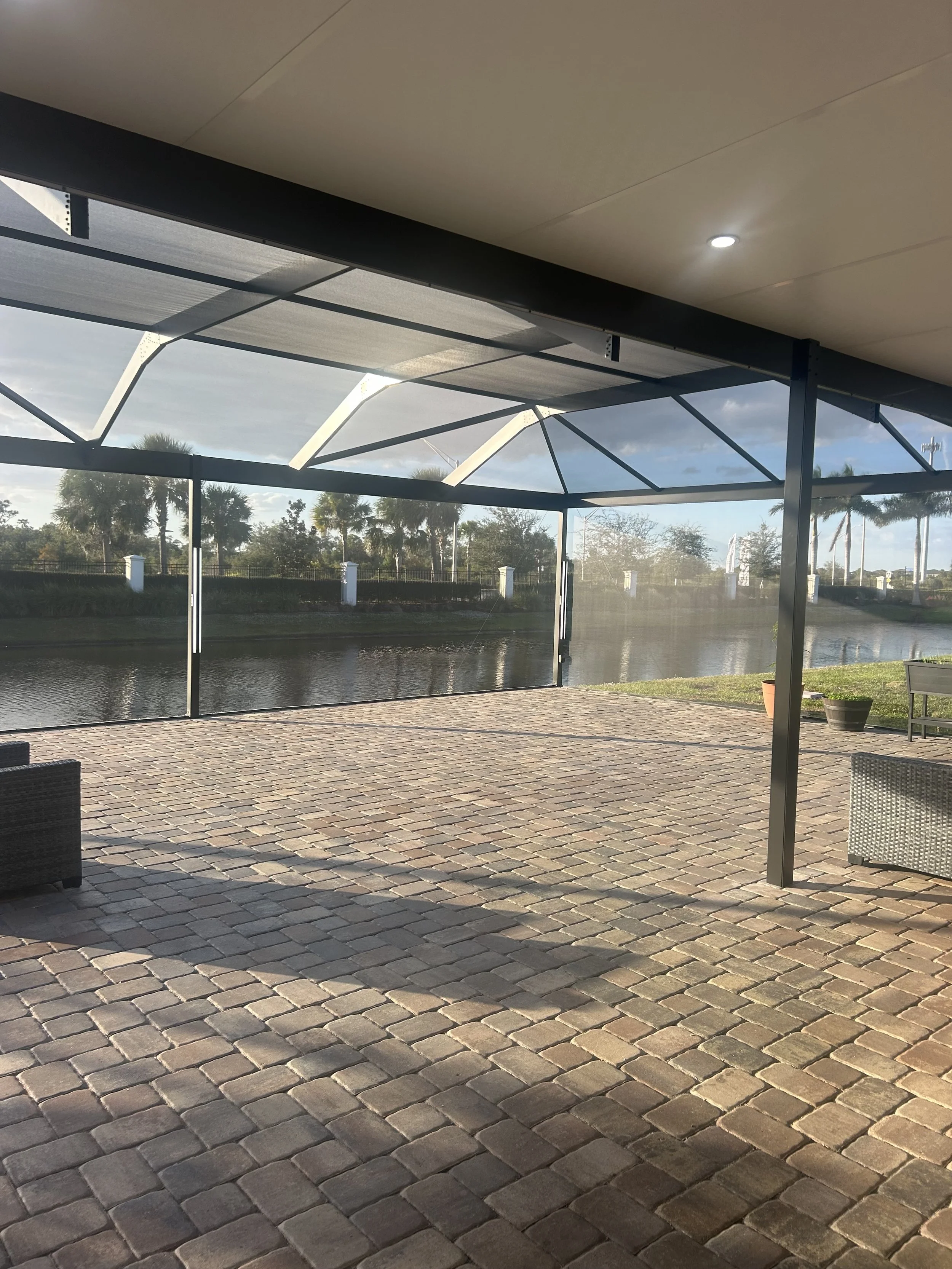 An outdoor patio with brick pavers, partially covered by a metal and fabric screened enclosure, overlooking a waterway with palm trees and blue sky in the background.