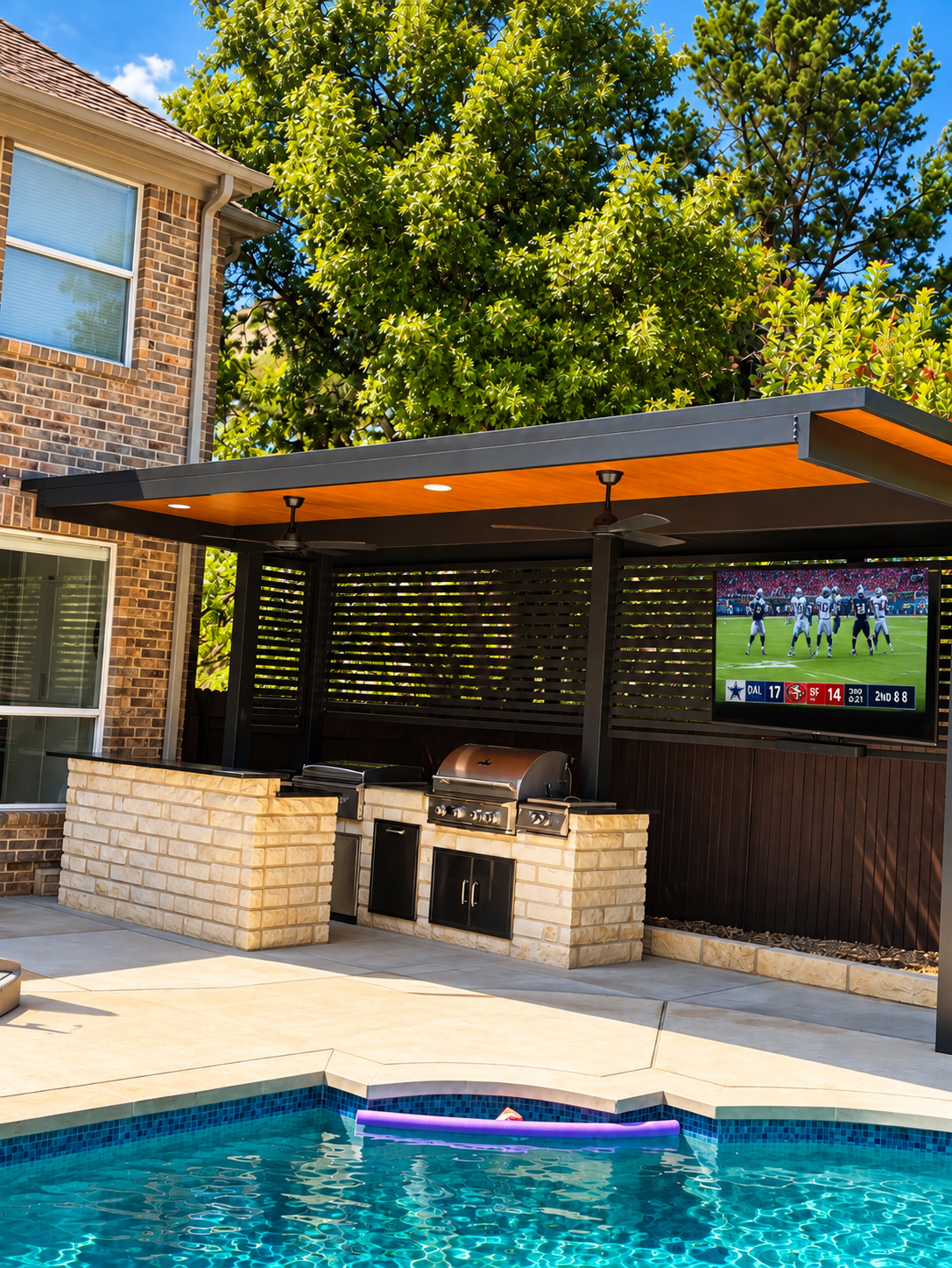 Backyard patio with swimming pool, outdoor kitchen with grill, television showing a football game, and large trees in the background.