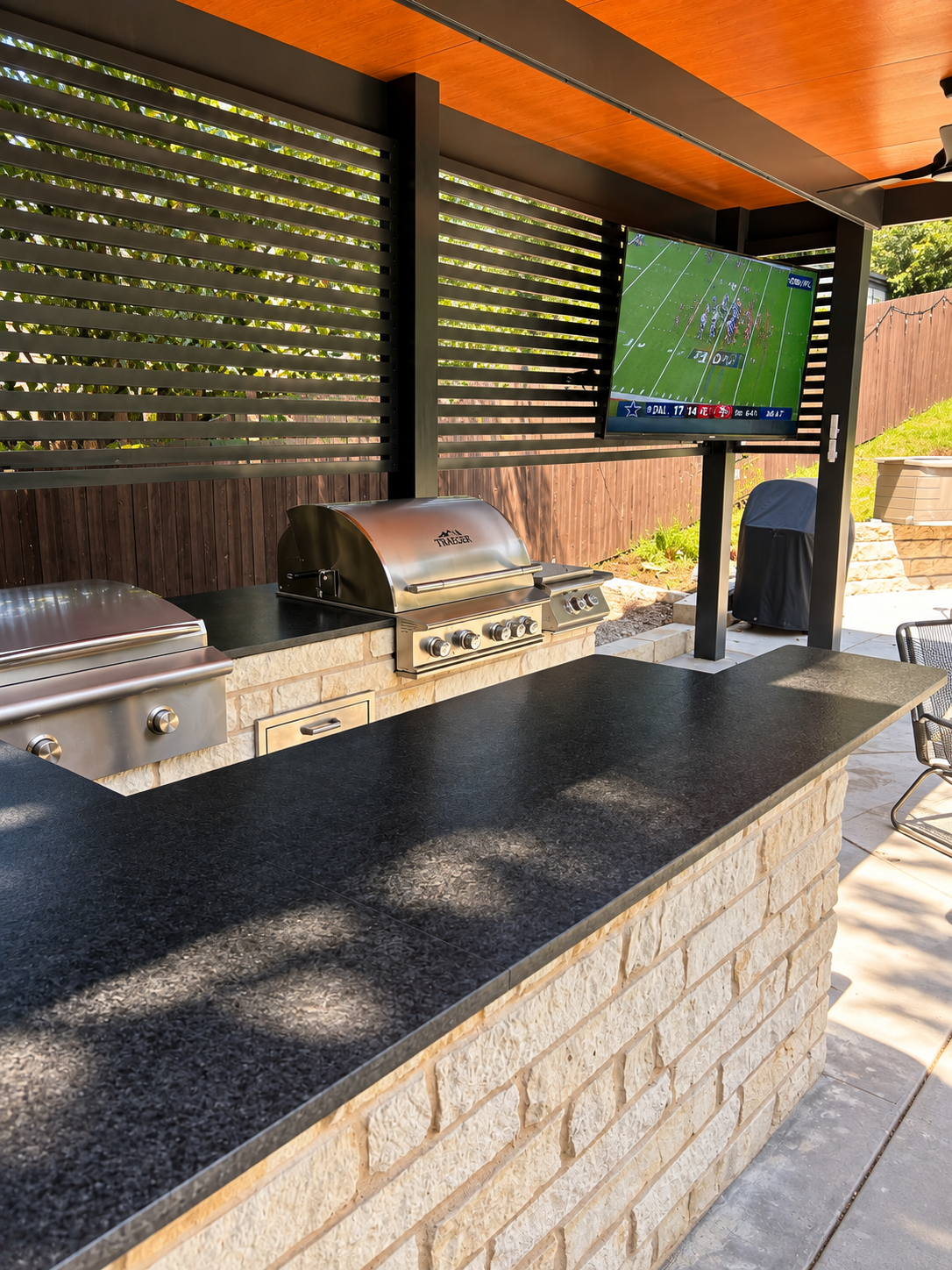 Outdoor entertainment area with stainless steel grills, black countertop, television showing a football game, wooden ceiling, and privacy fencing.