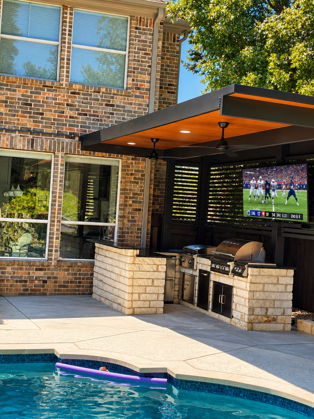 Backyard patio with swimming pool, outdoor kitchen with grill, television showing a football game, brick house wall, and trees.