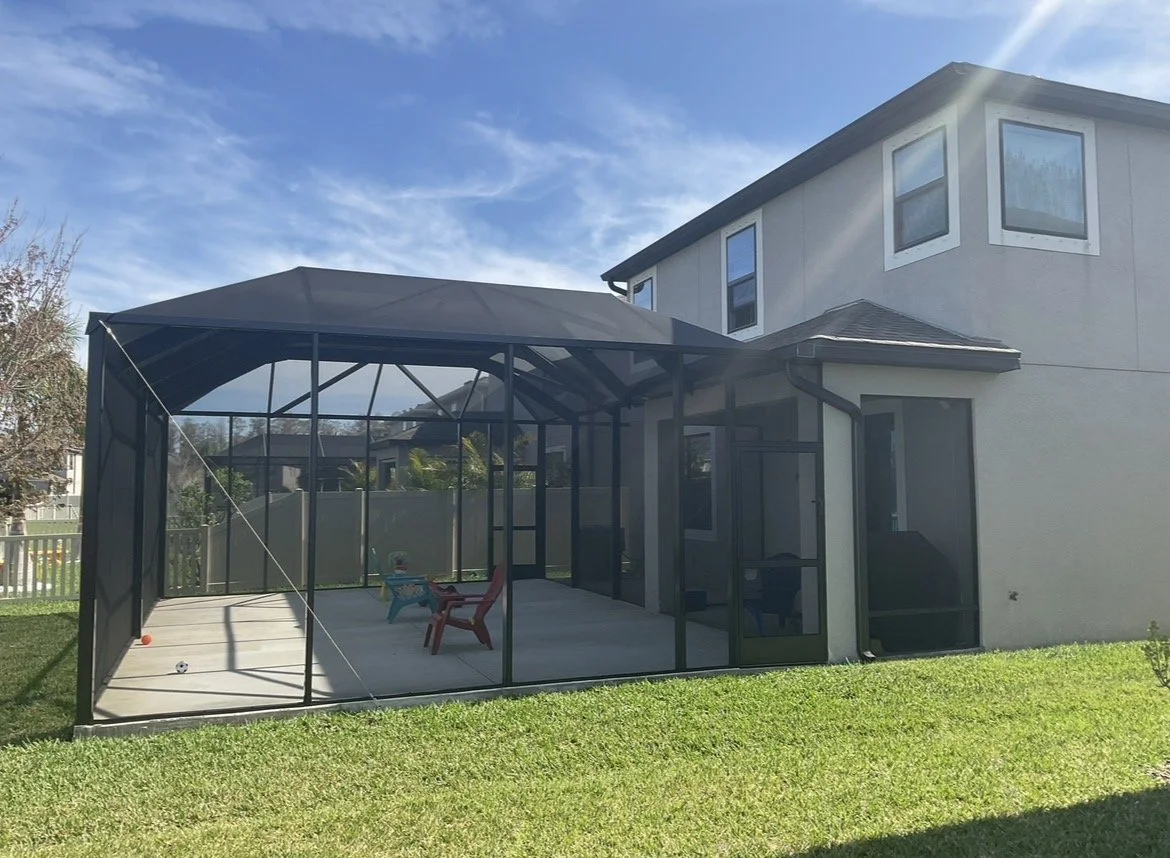Backyard enclosed patio with screened walls and a roof, with chairs and a toy on the concrete floor, next to a two-story house.