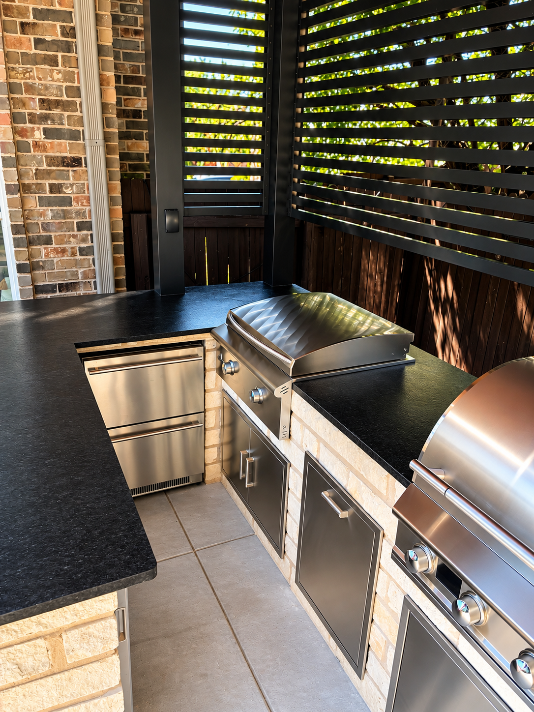 Outdoor kitchen with stainless steel grill and cabinets, black countertop, brick and wooden wall, black slatted privacy screen, and tiled floor