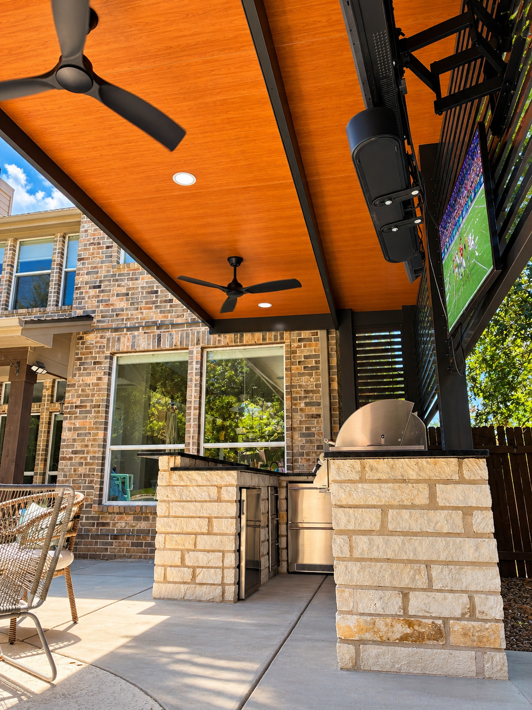 Outdoor patio with a ceiling fan, mounted TV showing a football game, and a built-in grill, against brick walls and a wooden ceiling.