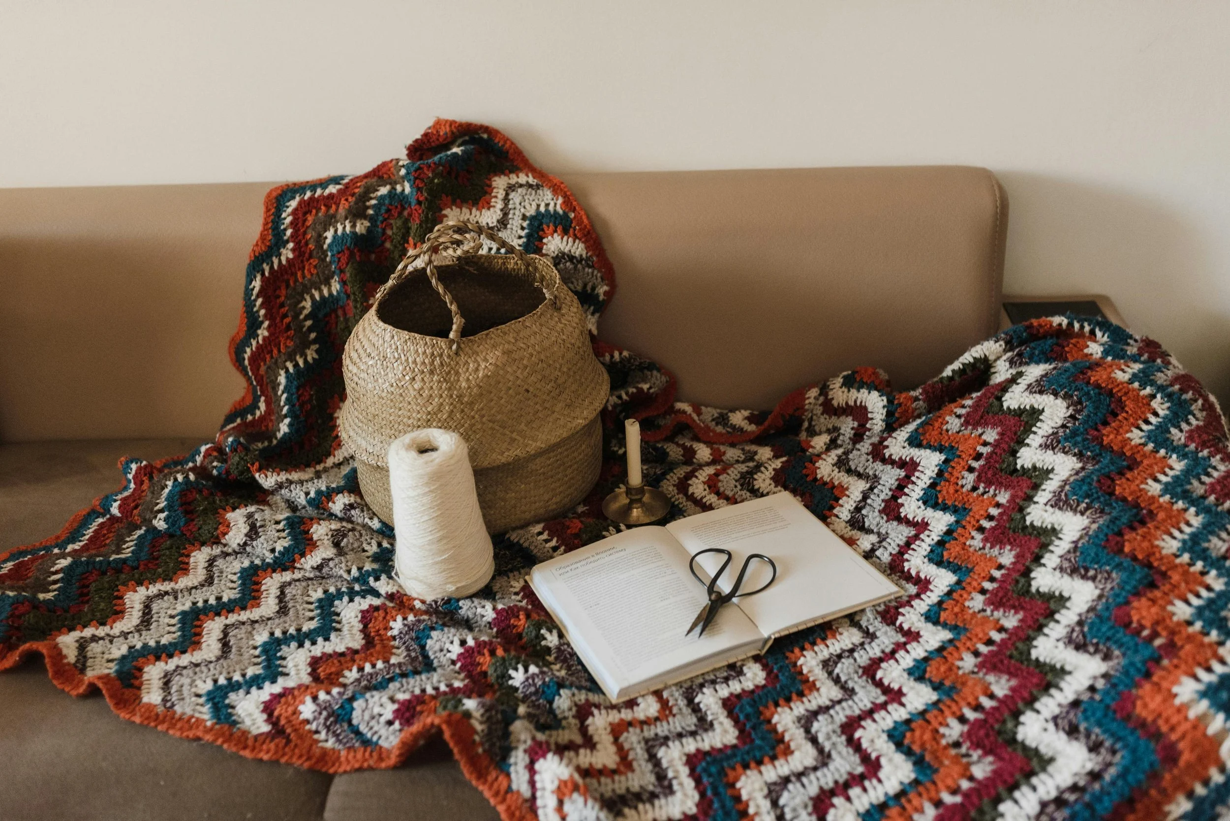 A cozy setup with a knitted blanket, a woven bag, a spool of white thread, a candle on a stand, an open book with scissors on top, and a beige headboard against a plain wall.