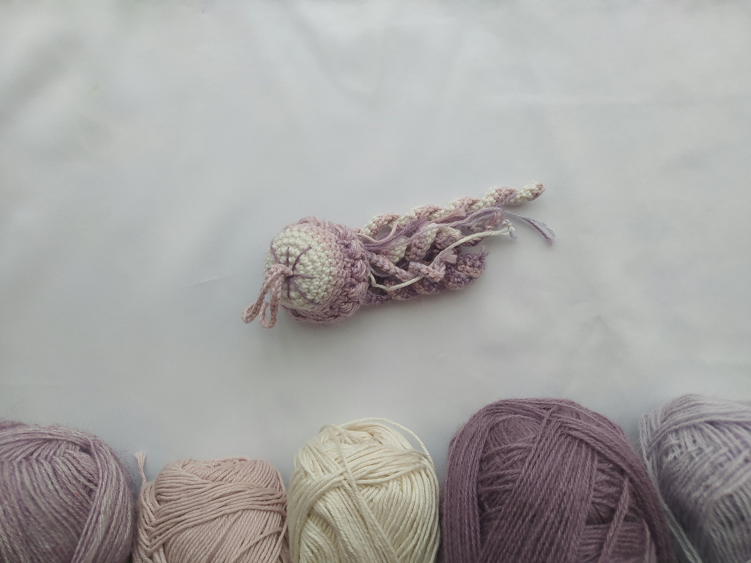 A small handmade crochet octopus toy in shades of pink and white, surrounded by balls of yarn in similar colors, on a plain white surface.