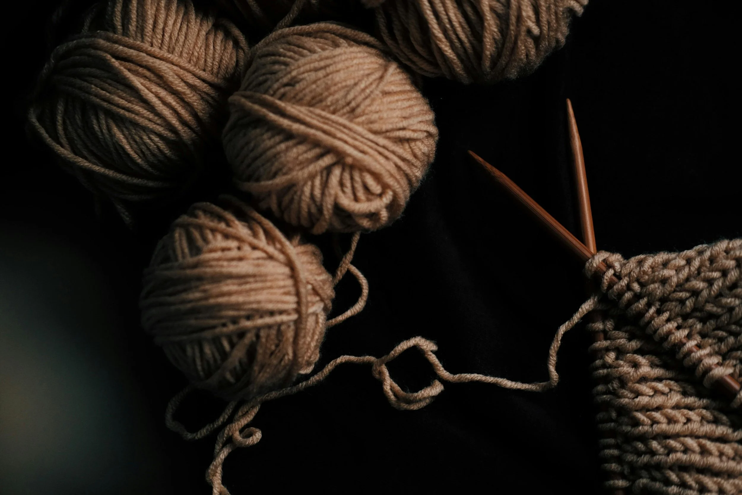 Balls of beige yarn, knitting needles, and a partially knitted piece of fabric on a dark background.