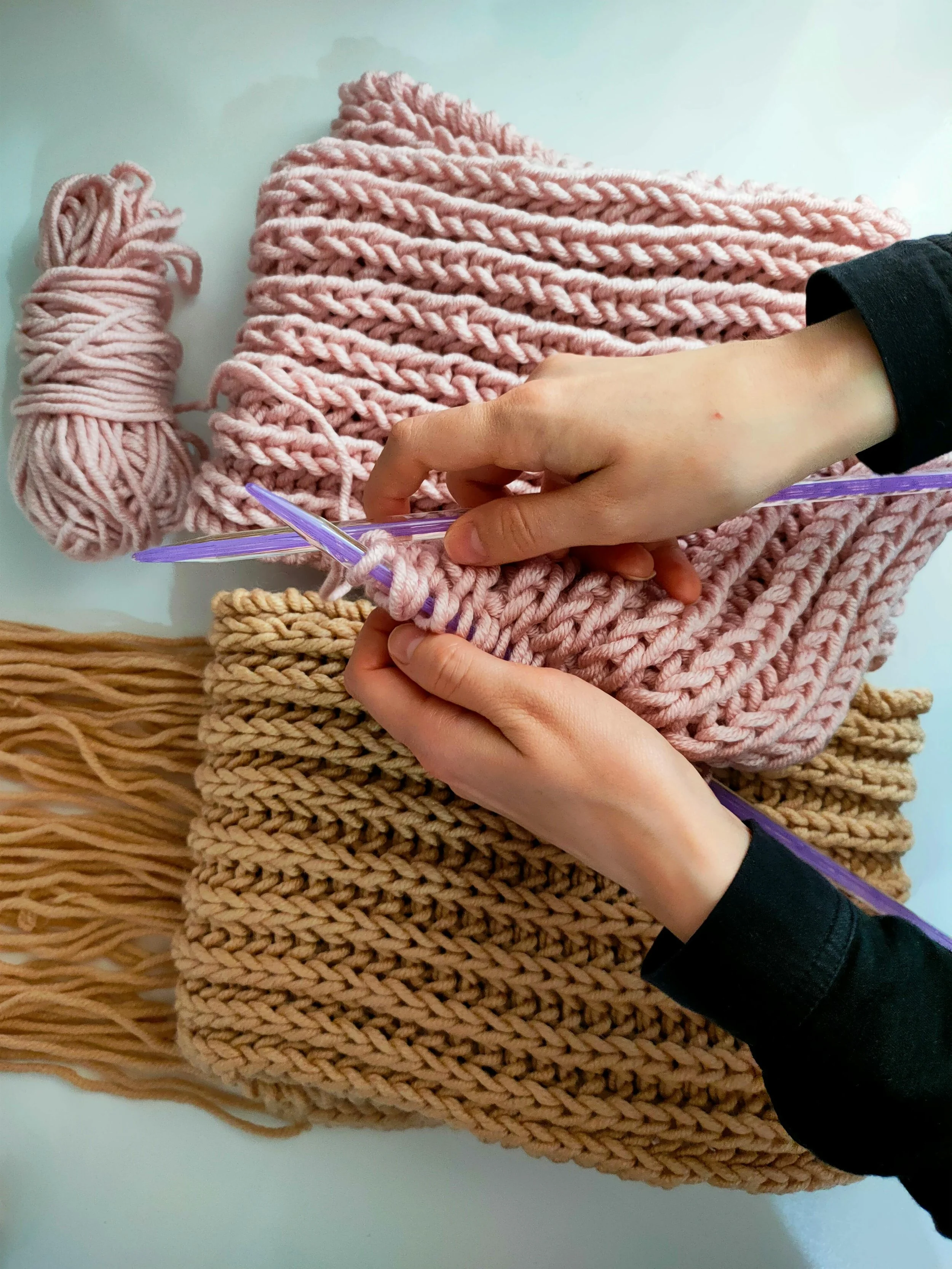 Person knitting pink chunky yarn with purple knitting needles, surrounded by finished knitting projects in pink and beige.