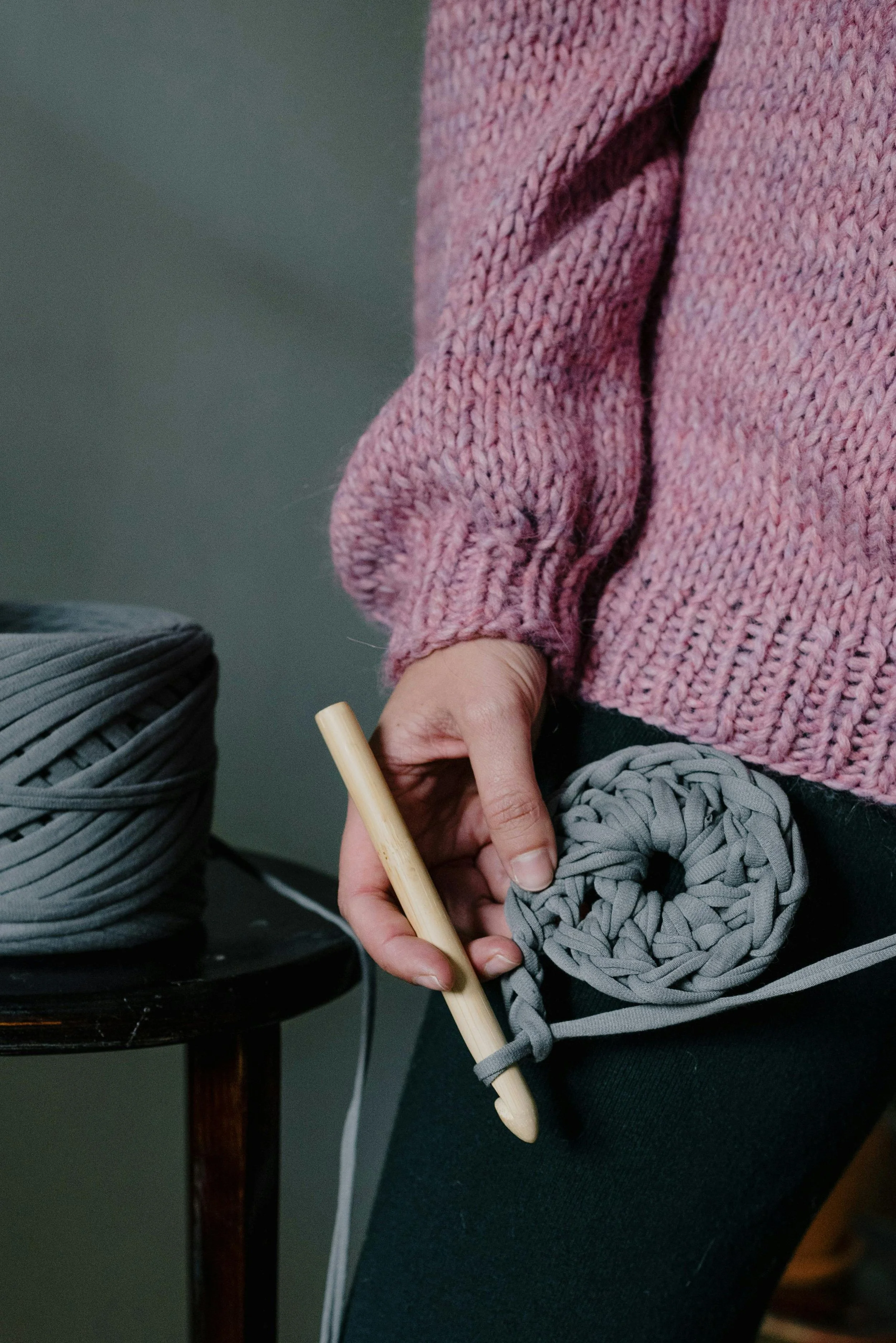 Close-up of a person knitting with grey yarn and a wooden crochet hook, wearing a pink chunky sweater.