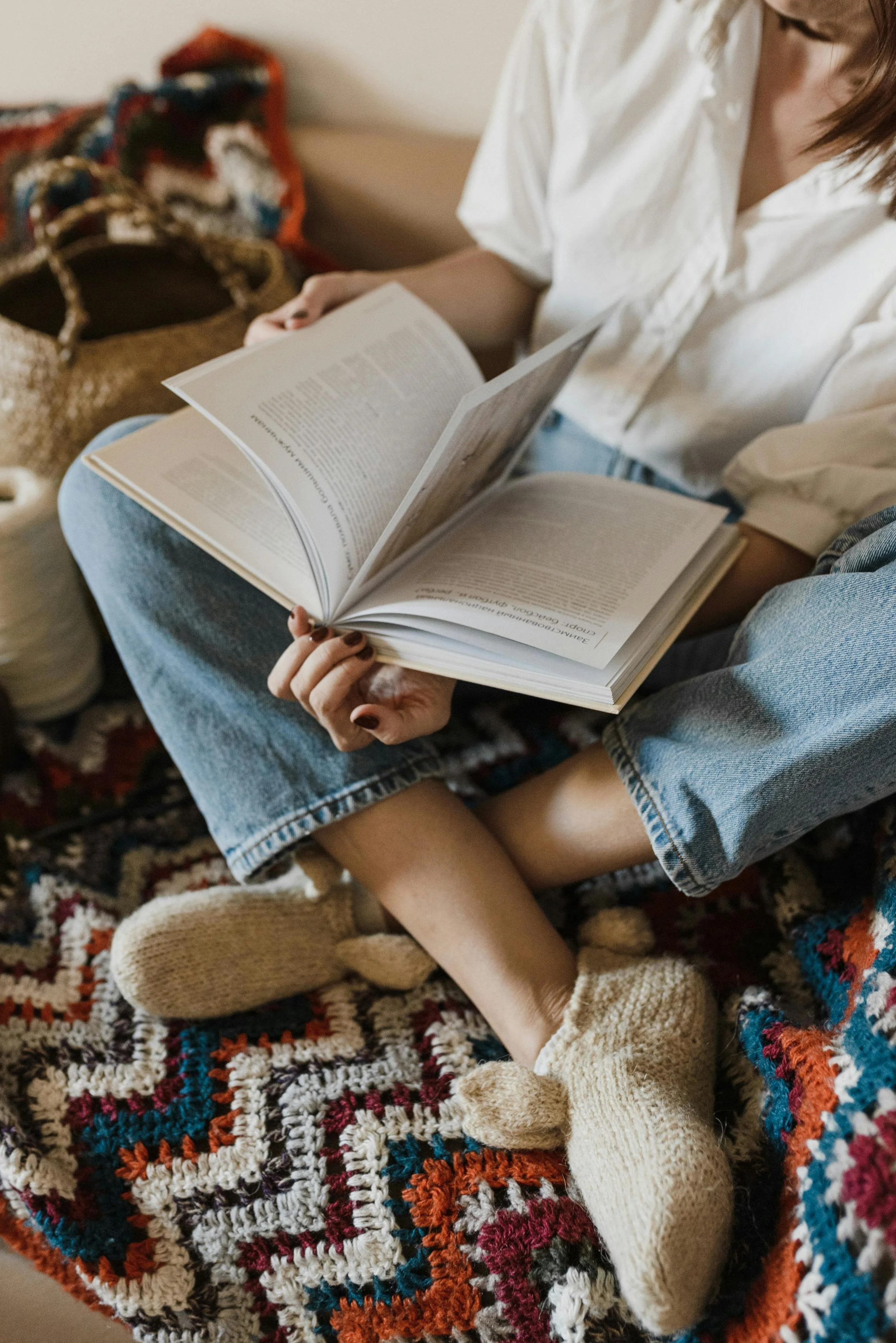 Person sitting cross-legged on a colorful crocheted blanket, reading a large open book, with a woven bag and a patterned pillow in the background.