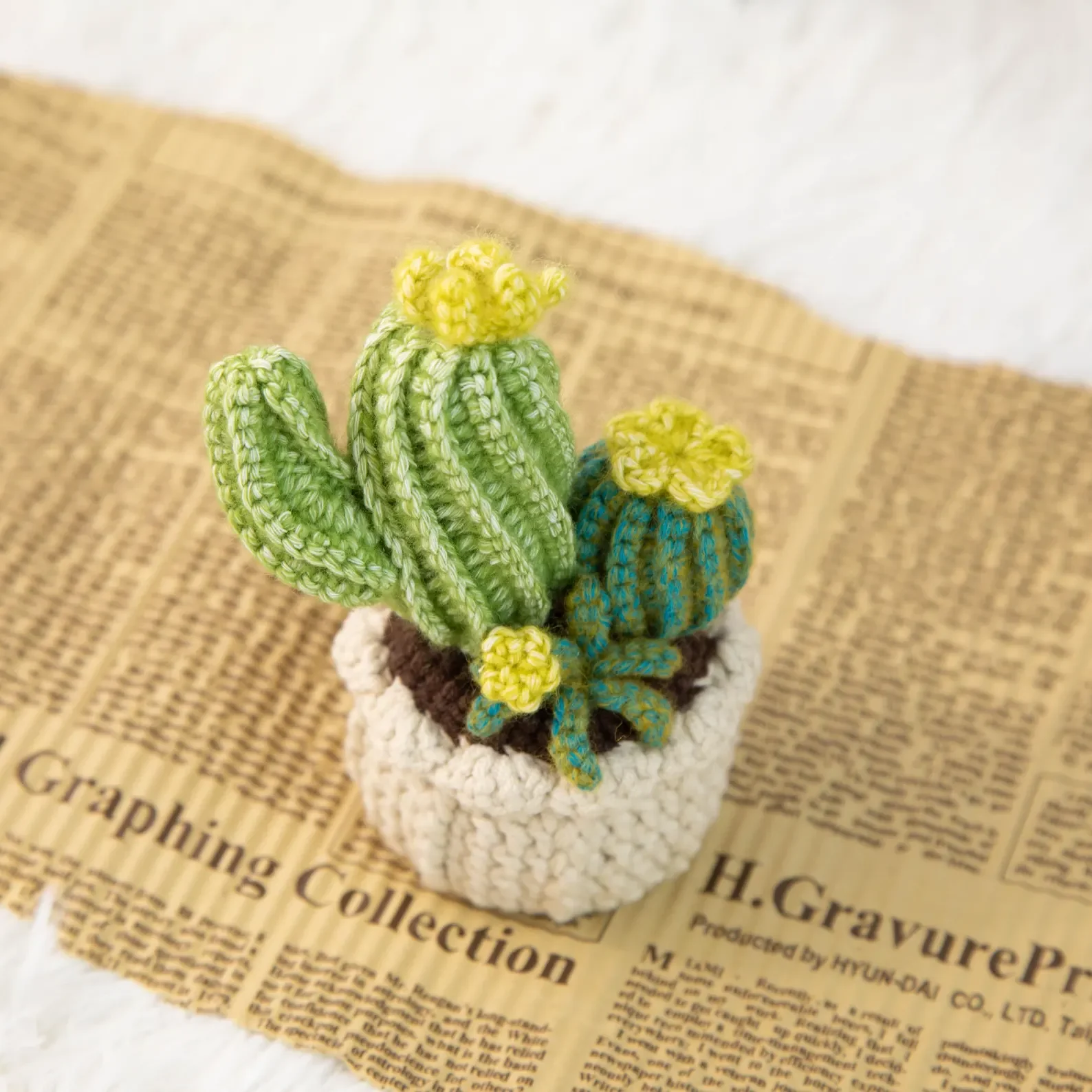Handmade crocheted cactus and succulent plants in a white pot placed on a sheet of newspaper.