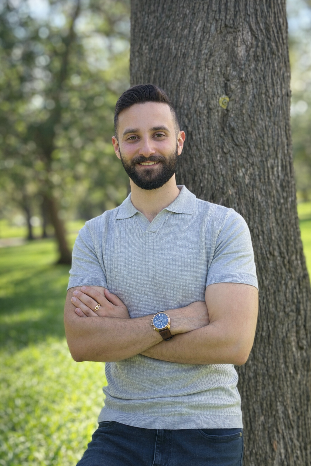 A man with a beard and short dark hair standing outdoors next to a tree, smiling with arms crossed, wearing a light gray textured polo shirt, a watch on his left wrist, and a ring on his right hand.